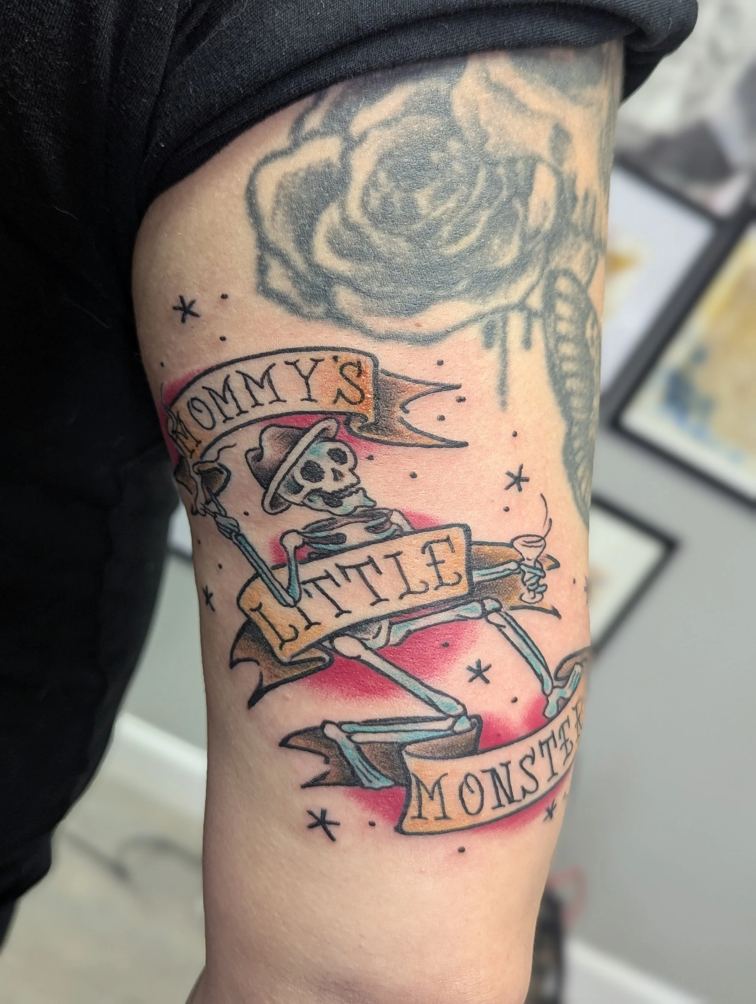 Colorful tattoo of a skeleton with a banner that reads 'mommy's little Monster' on a person's upper arm.