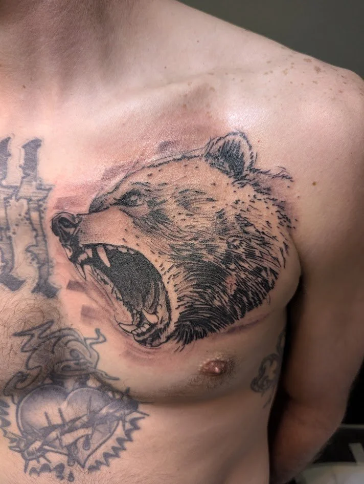 Tattoo of a roaring bear's head on a man's upper chest near the shoulder.