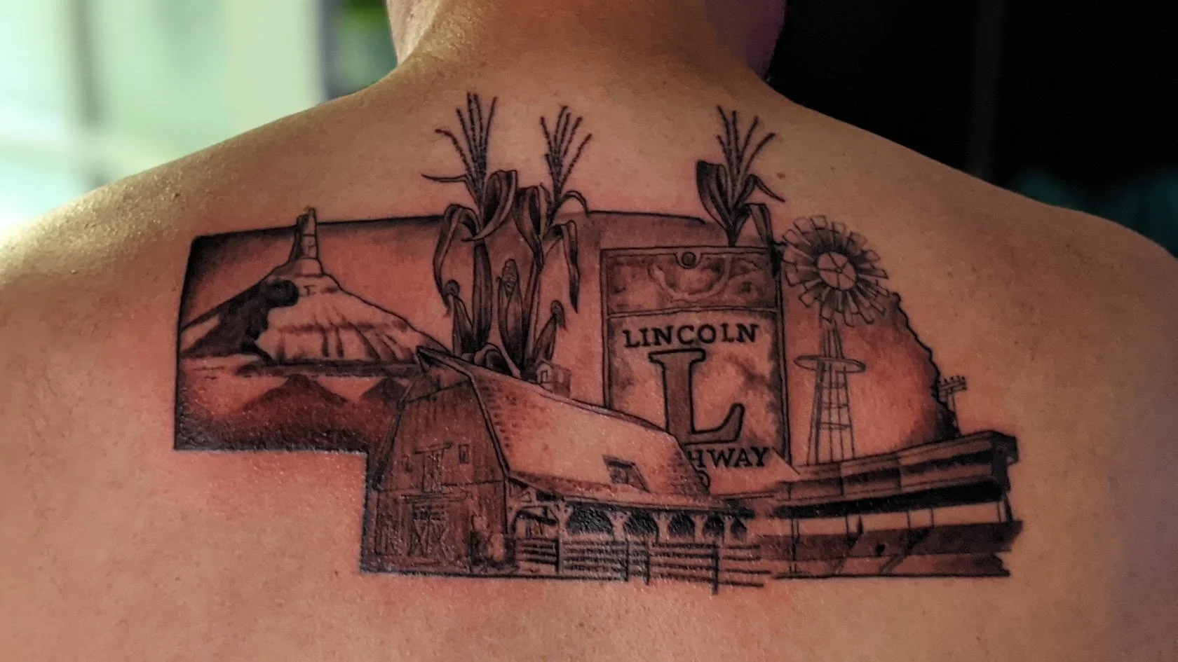 Tattoo of a landscape in Lincoln Highway, featuring a barn, windmill, and wheat stalks.
