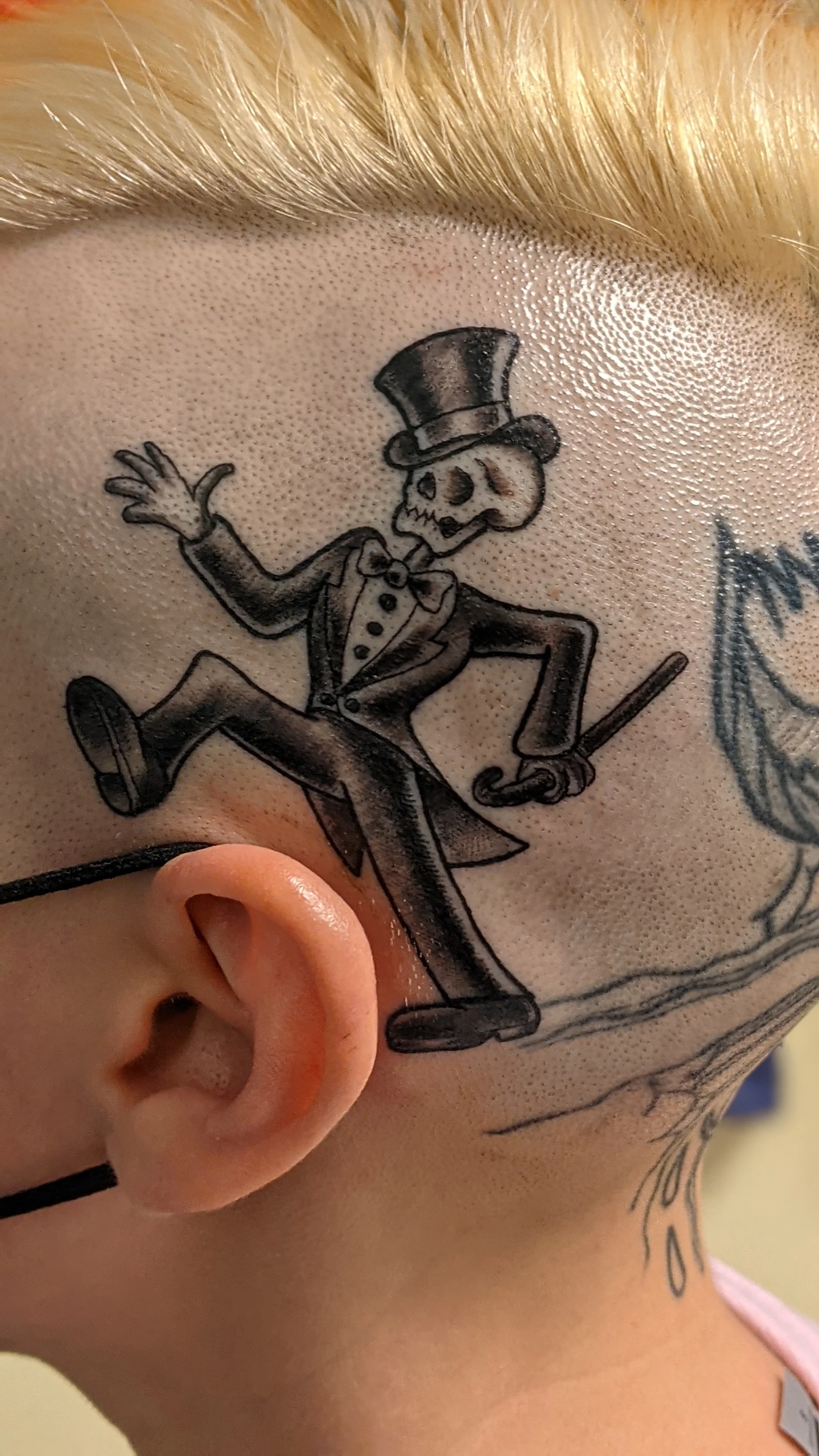 Tattoo of a skeleton wearing a top hat, suit, and bow tie, walking and waving, on a person's neck.