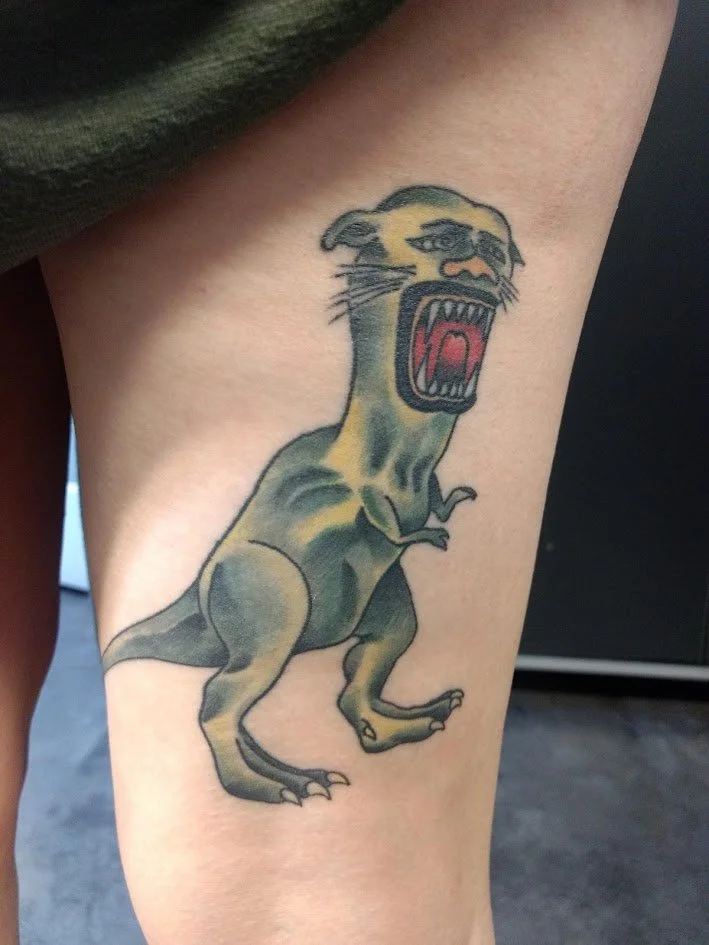 Tattoo of a snarling creature with a lion's body and a panther's face, showing sharp teeth and an open mouth, on a person's thigh.