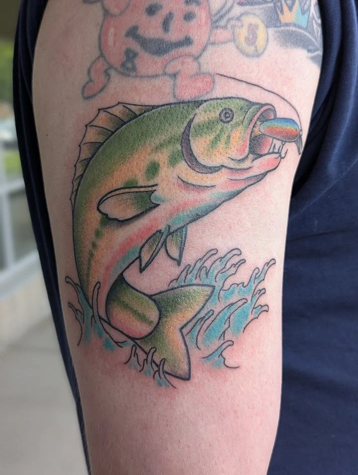 Colorful tattoo of a fish leaping out of water, with waves below and a small part of a cartoon character above.