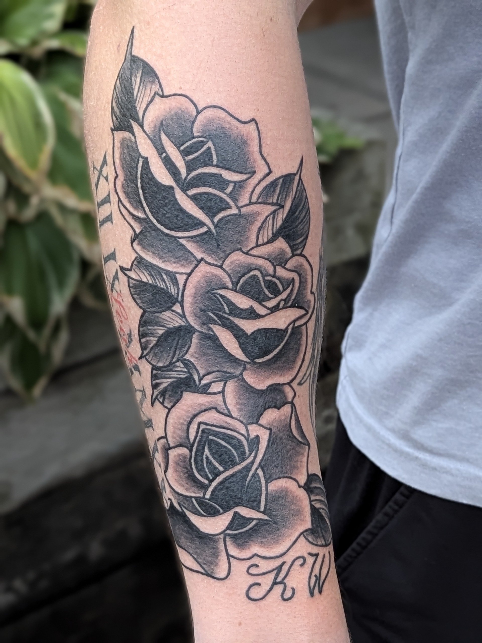 Close-up of a person's arm with a black and gray tattoo of roses and leaves, and cursive writing near the wrist.