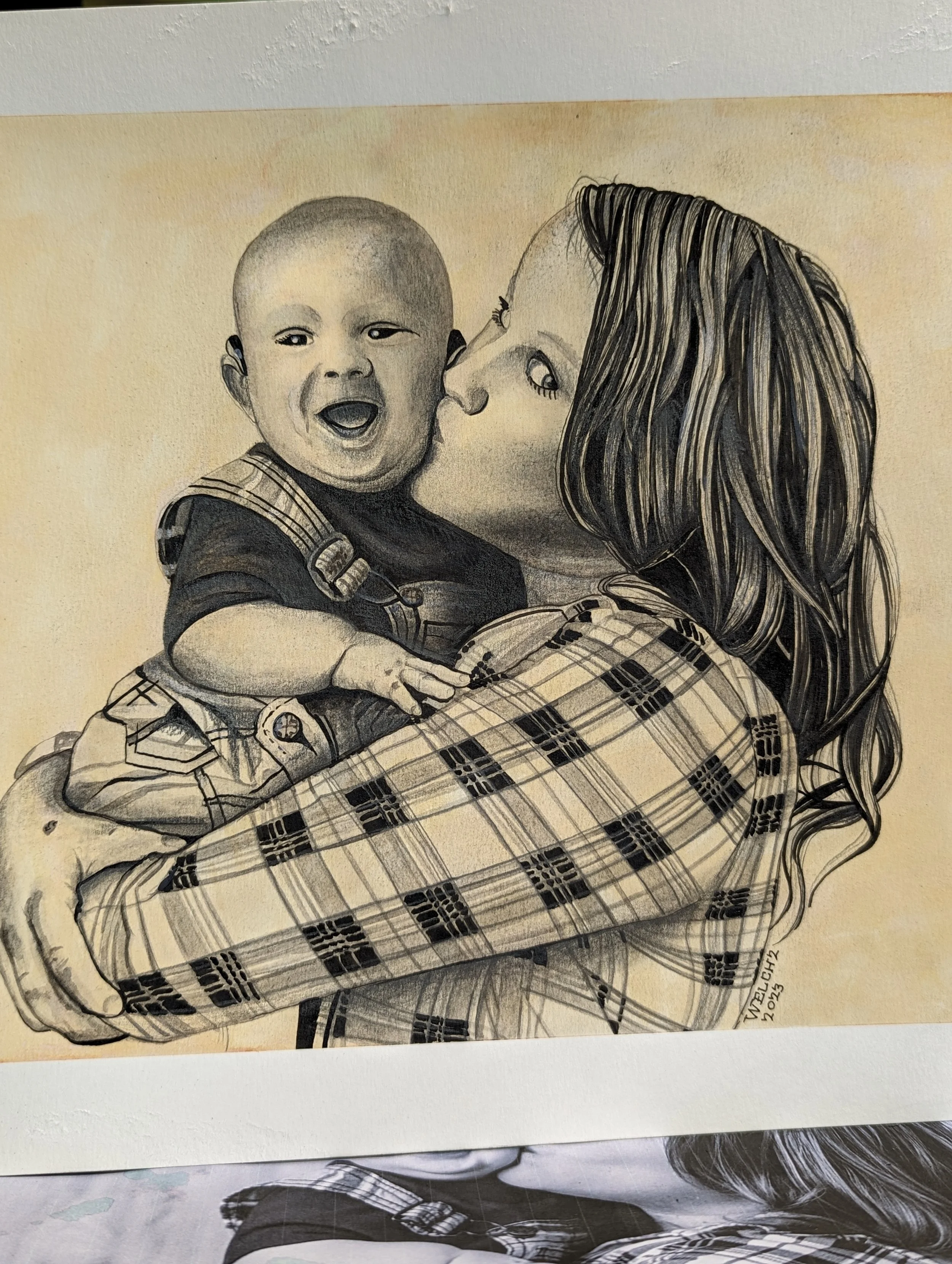 A black and white drawing of a woman holding a young child, who is smiling and looking at the woman, with the woman giving a gentle kiss on the child's face, wearing a plaid shirt.
