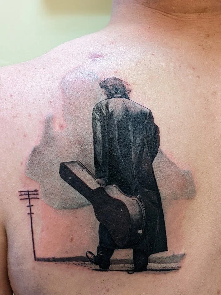 Tattoo of a person dressed in black, walking away and carrying a guitar case, on a person's chest.