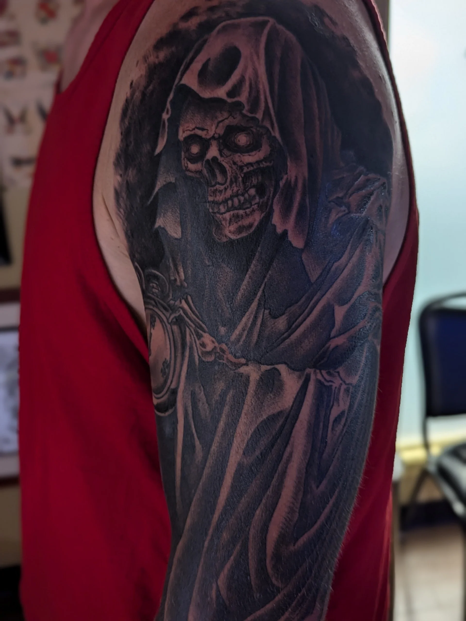 A close-up of a tattoo on a person's upper arm, featuring a detailed image of a skull with a hood, rendered in black ink.