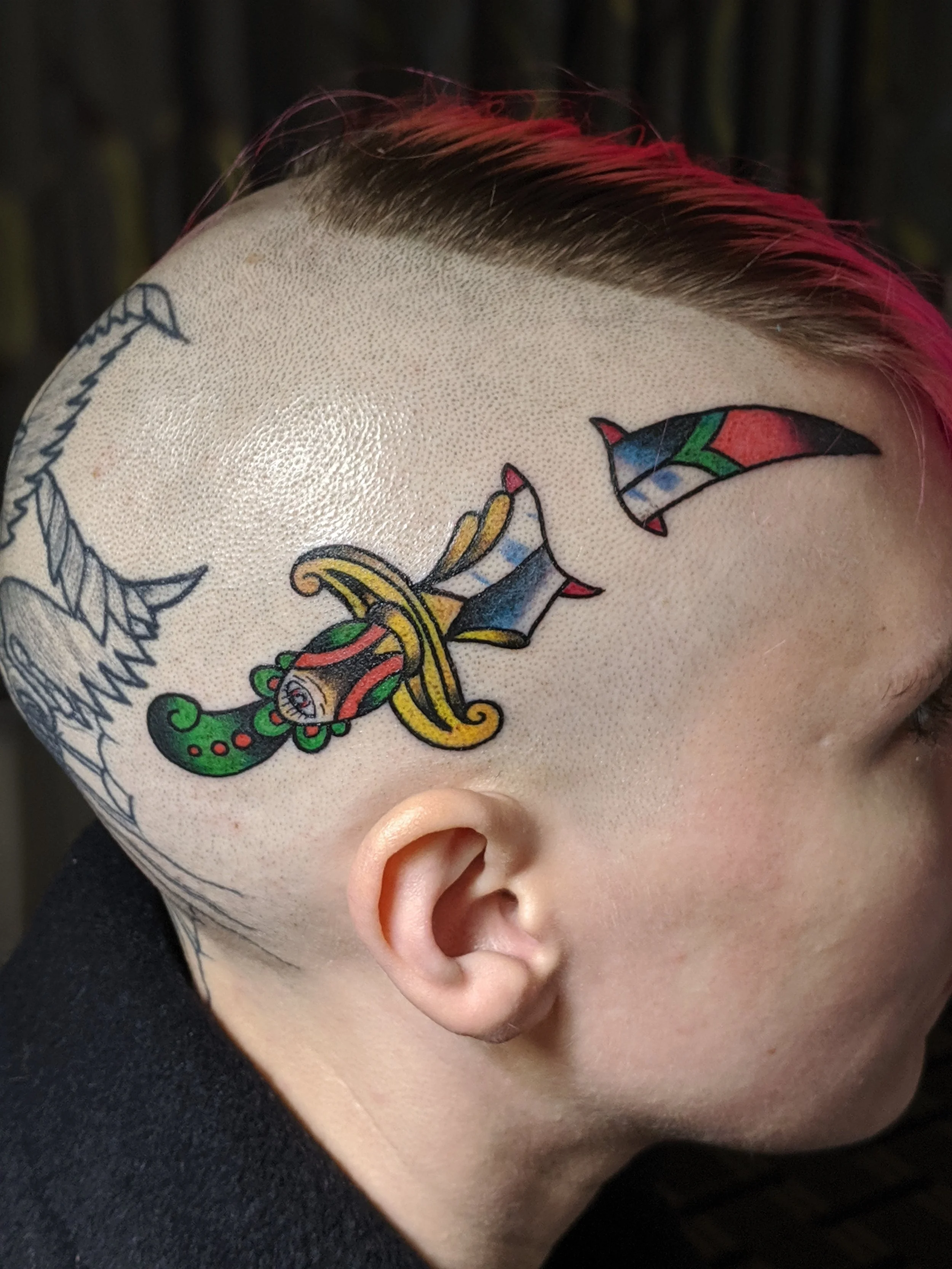 Close-up of a person's face with a colorful tattoo around the ear, featuring a snake design with green, yellow, red, and blue colors, and part of a tattooed face with a leaf on the forehead.