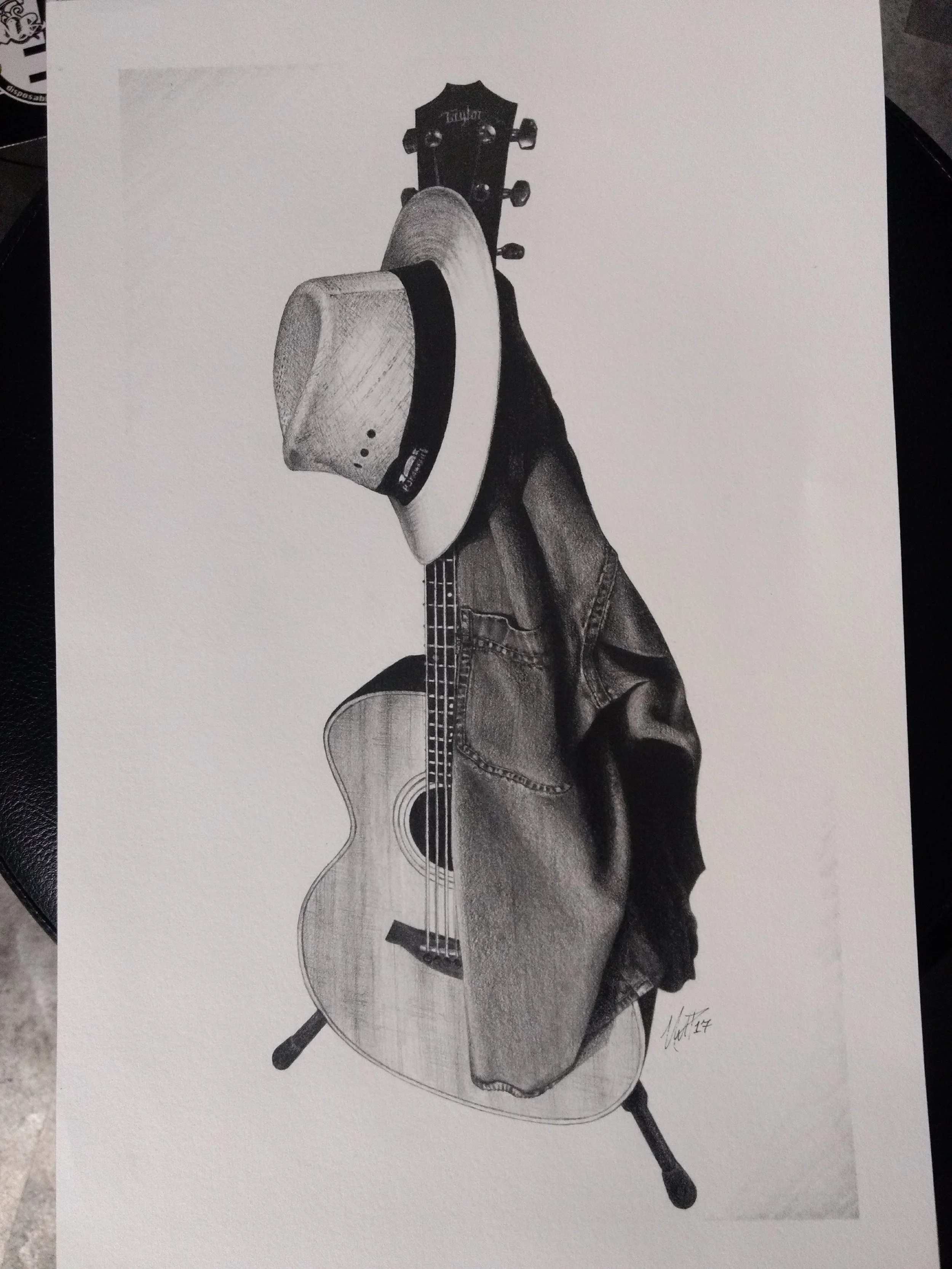 A detailed black and white drawing of an acoustic guitar with jeans-clad legs and feet, a hat hanging from the guitar's neck, and a small signature in the bottom right corner.