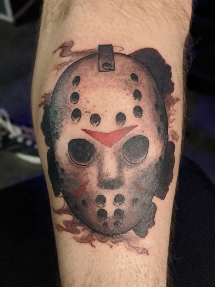 Tattoo of a horror movie mask with burnt effect, eye holes, red markings, and smoke surrounding it.