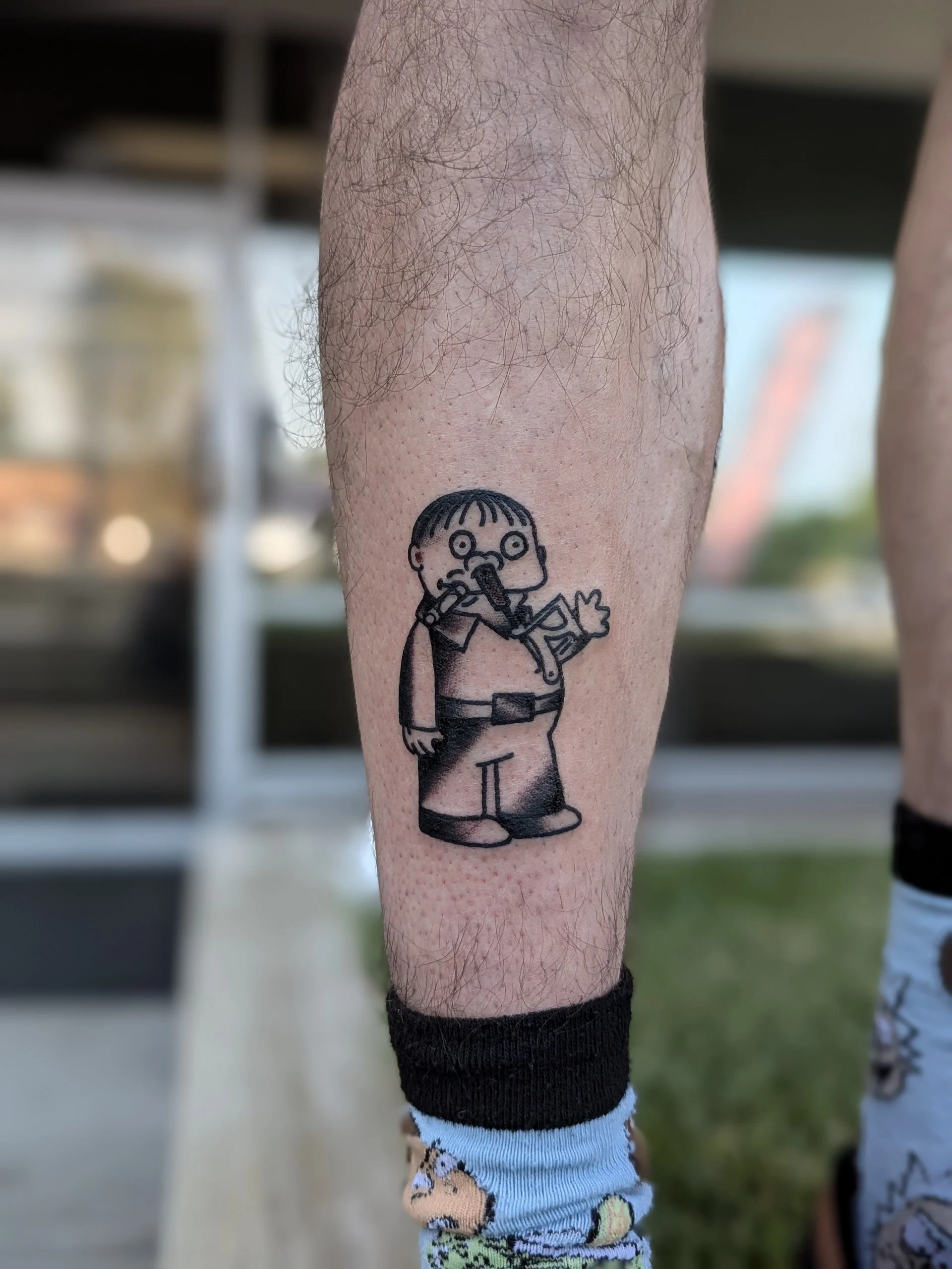 Close-up of a person's leg with a black outline tattoo of a cartoon character with glasses, holding a food item, and wearing shorts, with other legs and socks visible in the background.