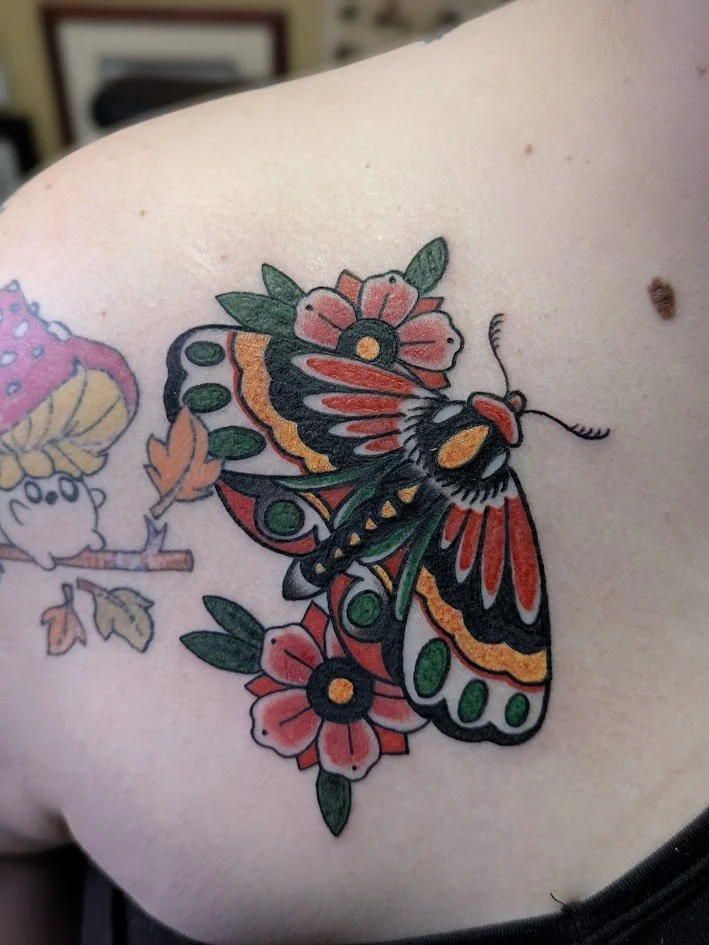 Colorful tattoo of a butterfly with red, black, green, and yellow patterns, surrounded by pink and red flowers with green leaves.