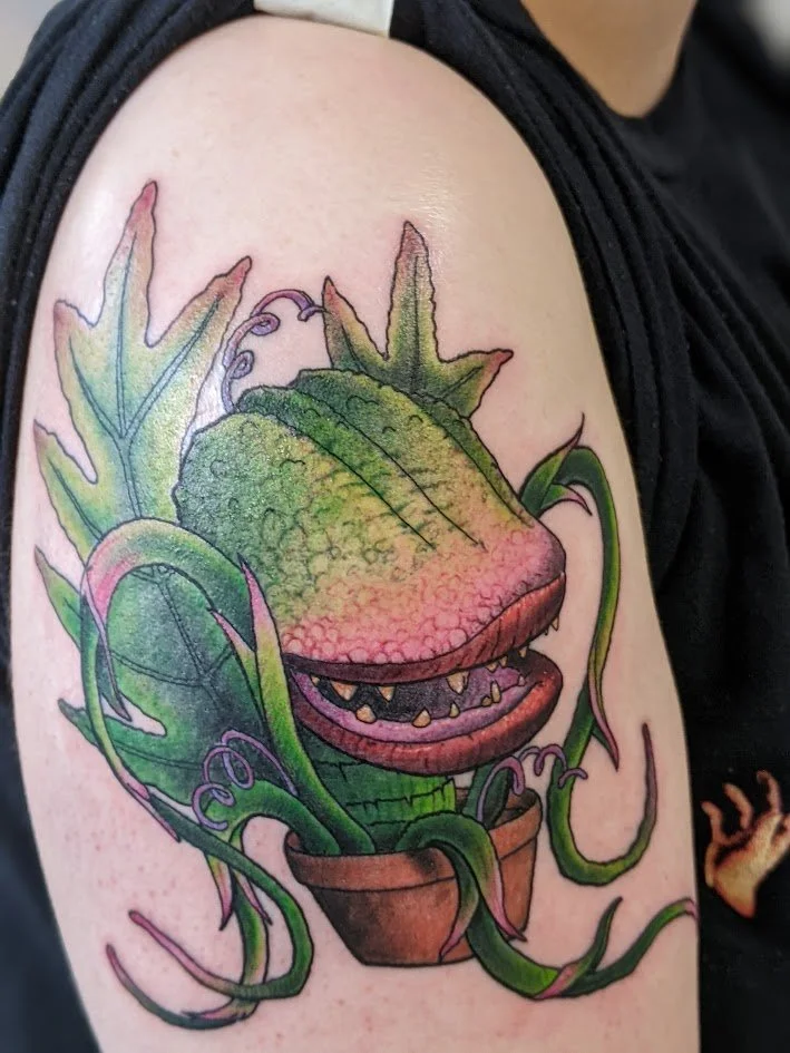 Tattoo of a potted plant with a dinosaur-like head, leafy green surroundings, and tentacle-like vines on the upper arm.