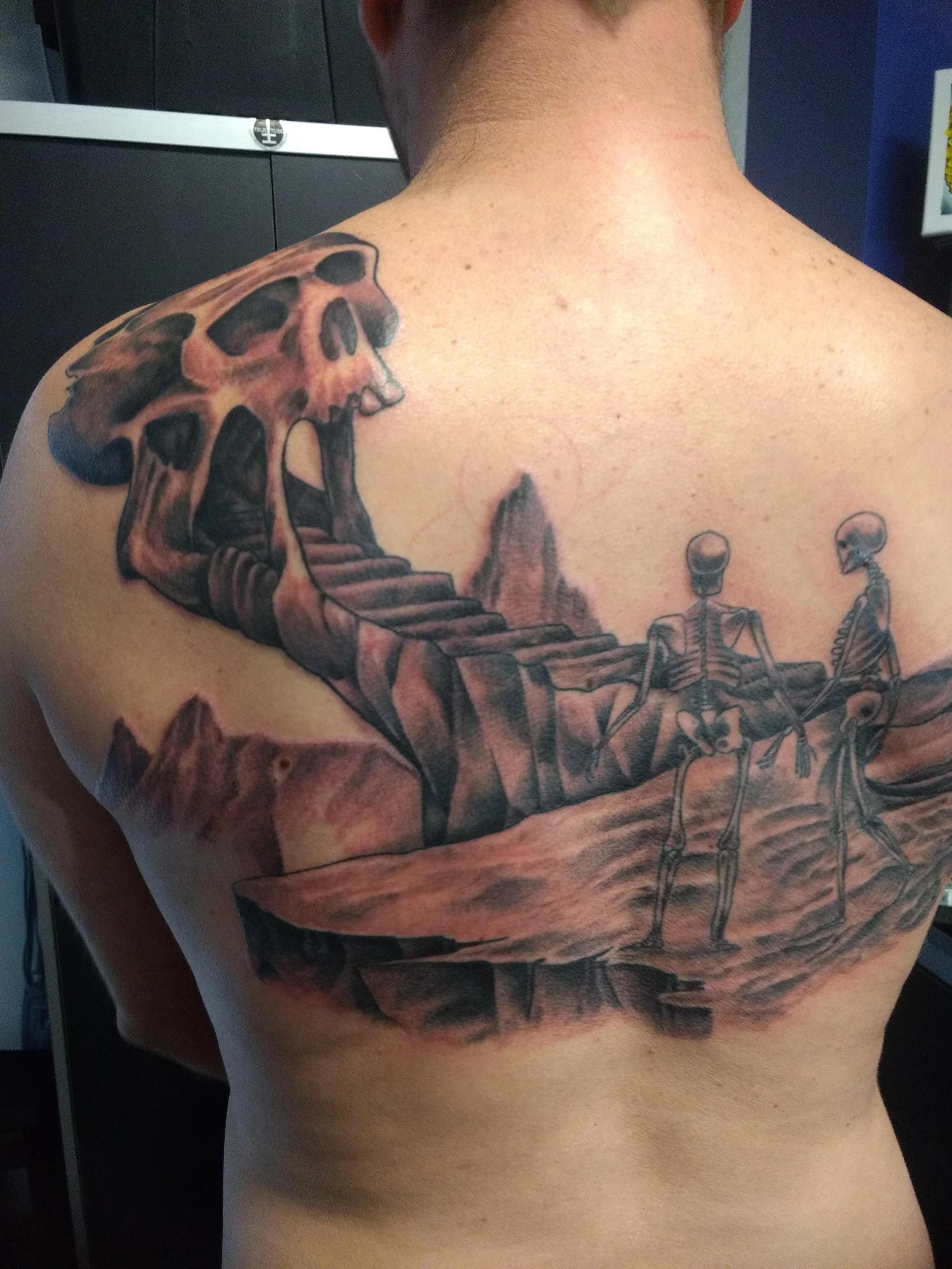 Tattoo on a person's shoulder and upper chest depicting a skeleton with stairs leading into its mouth, set against a surreal landscape with mountains and two smaller skeletons.