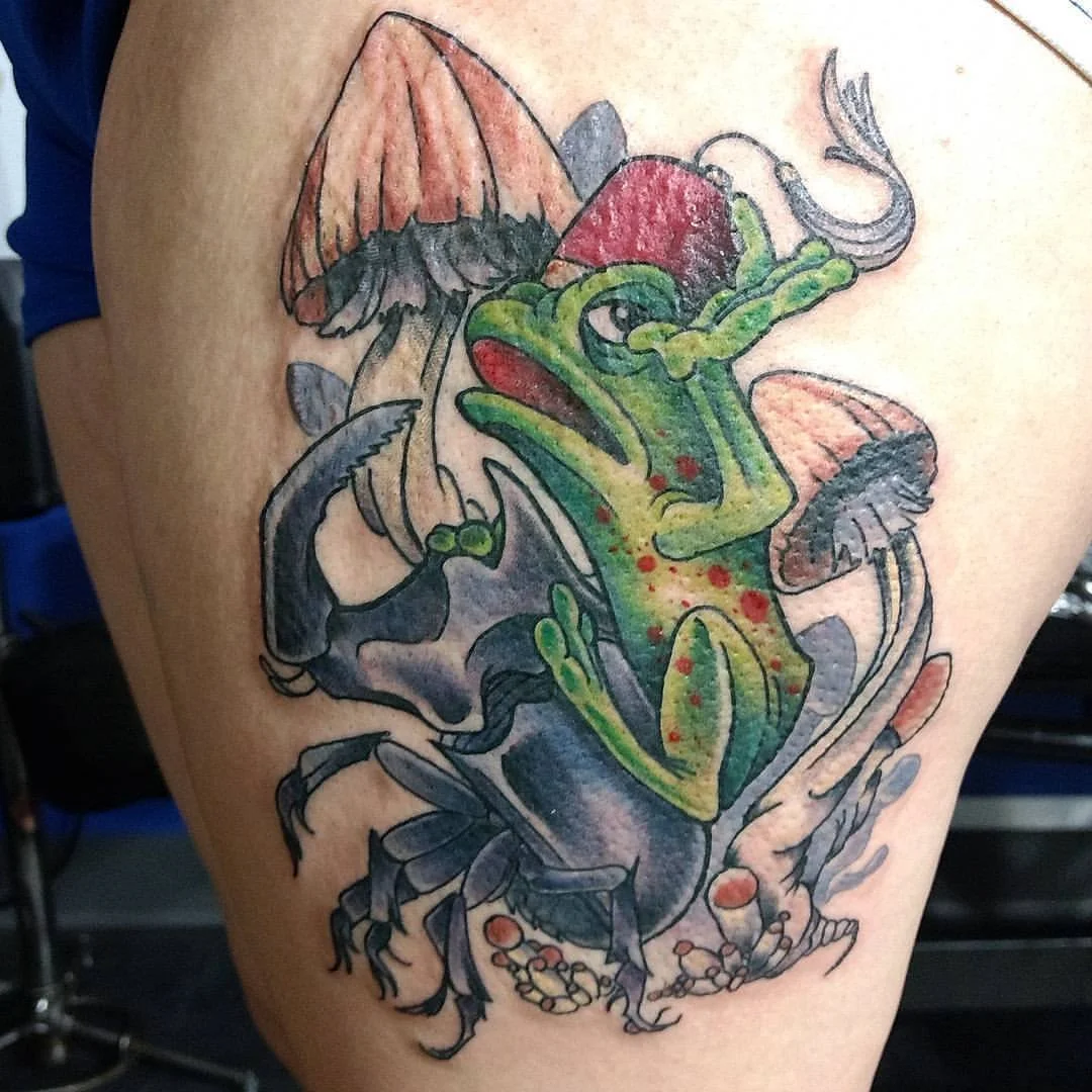 A tattoo depicting a green frog sitting on a beetle with mushroom and leaf elements surrounding them.