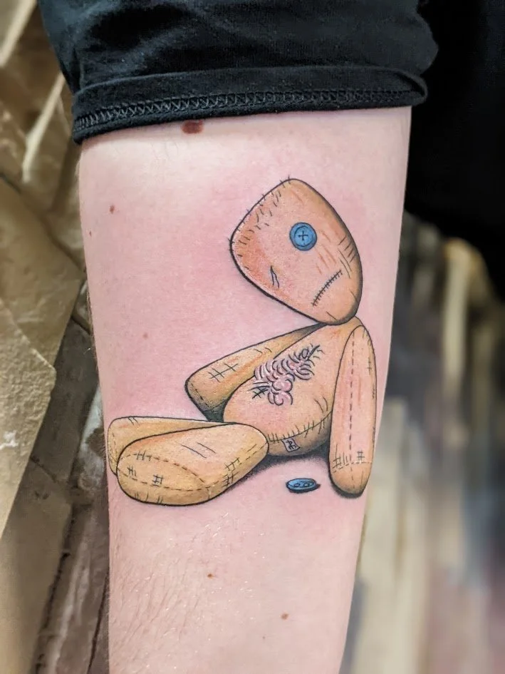 Tattoo of a stuffed teddy bear with a blue button eye, sitting with a small blue button nearby.