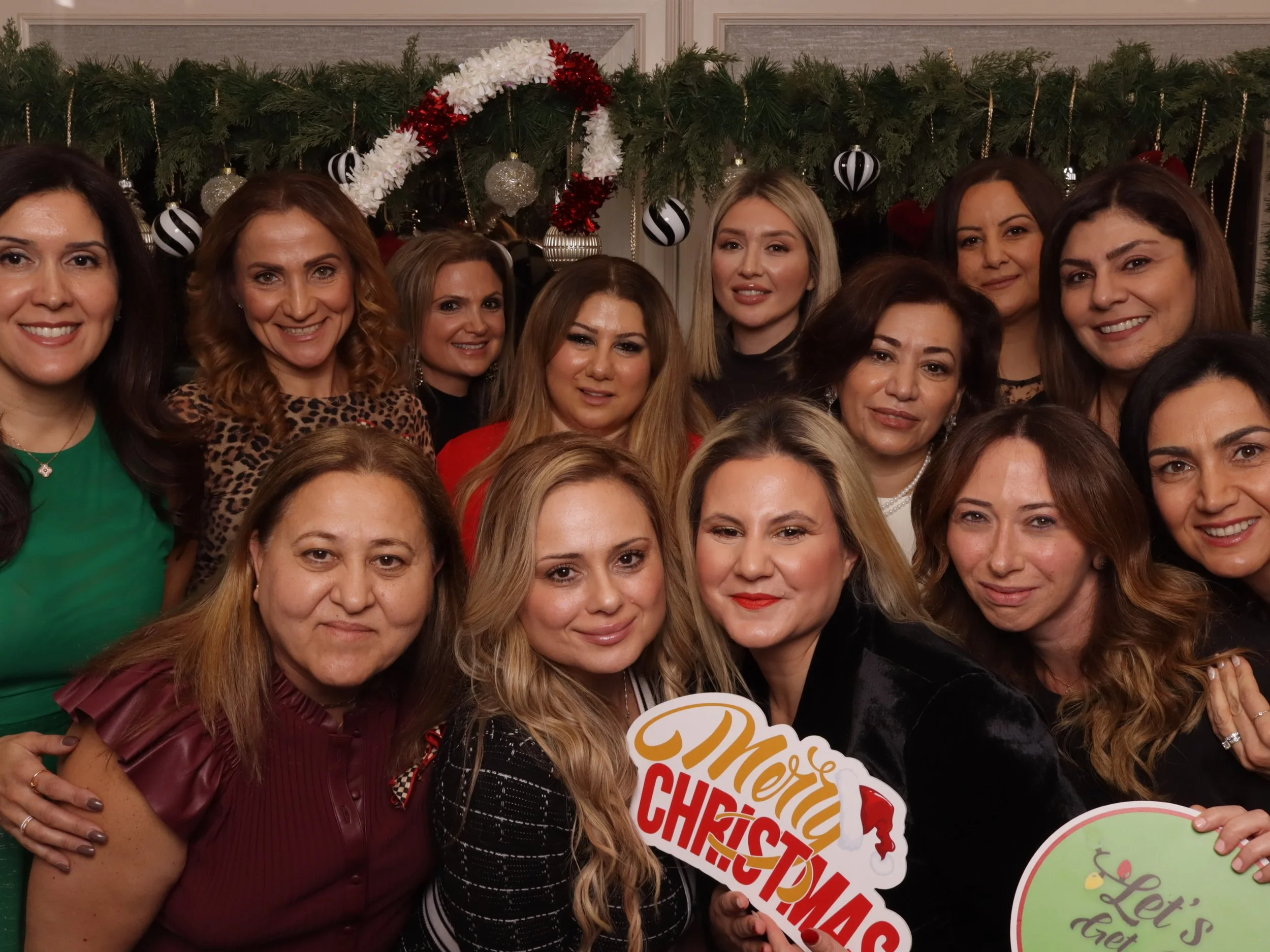 Group of women celebrating Christmas, standing in front of a decorated fireplace with a garland, ornaments, and holiday decorations, holding a sign that reads 'Merry Christmas' and a green sign with 'Let's Get' visible.