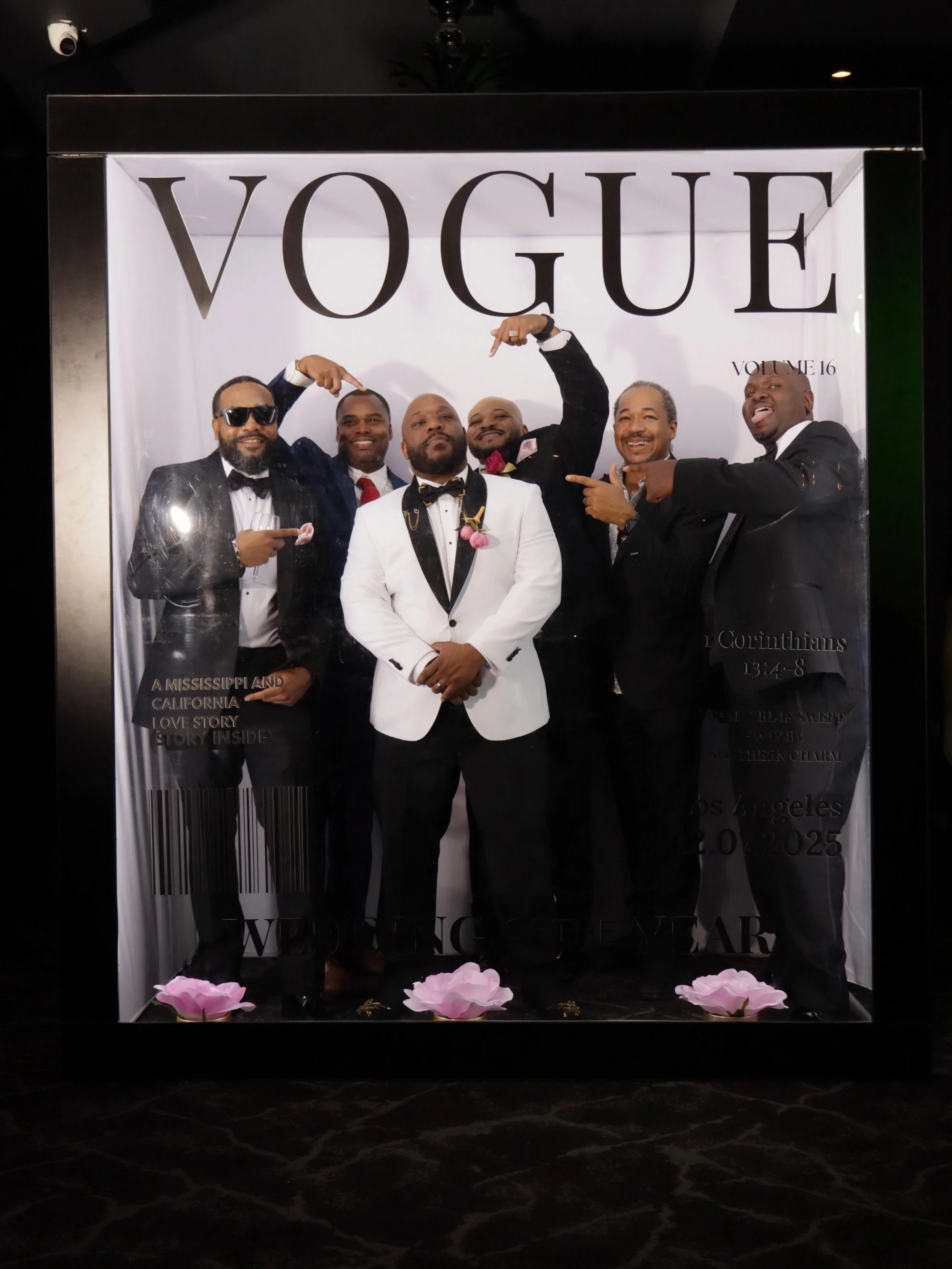 Group of six men in tuxedos posing in front of a Vogue photo backdrop, some pointing and smiling, with pink flowers on the floor.