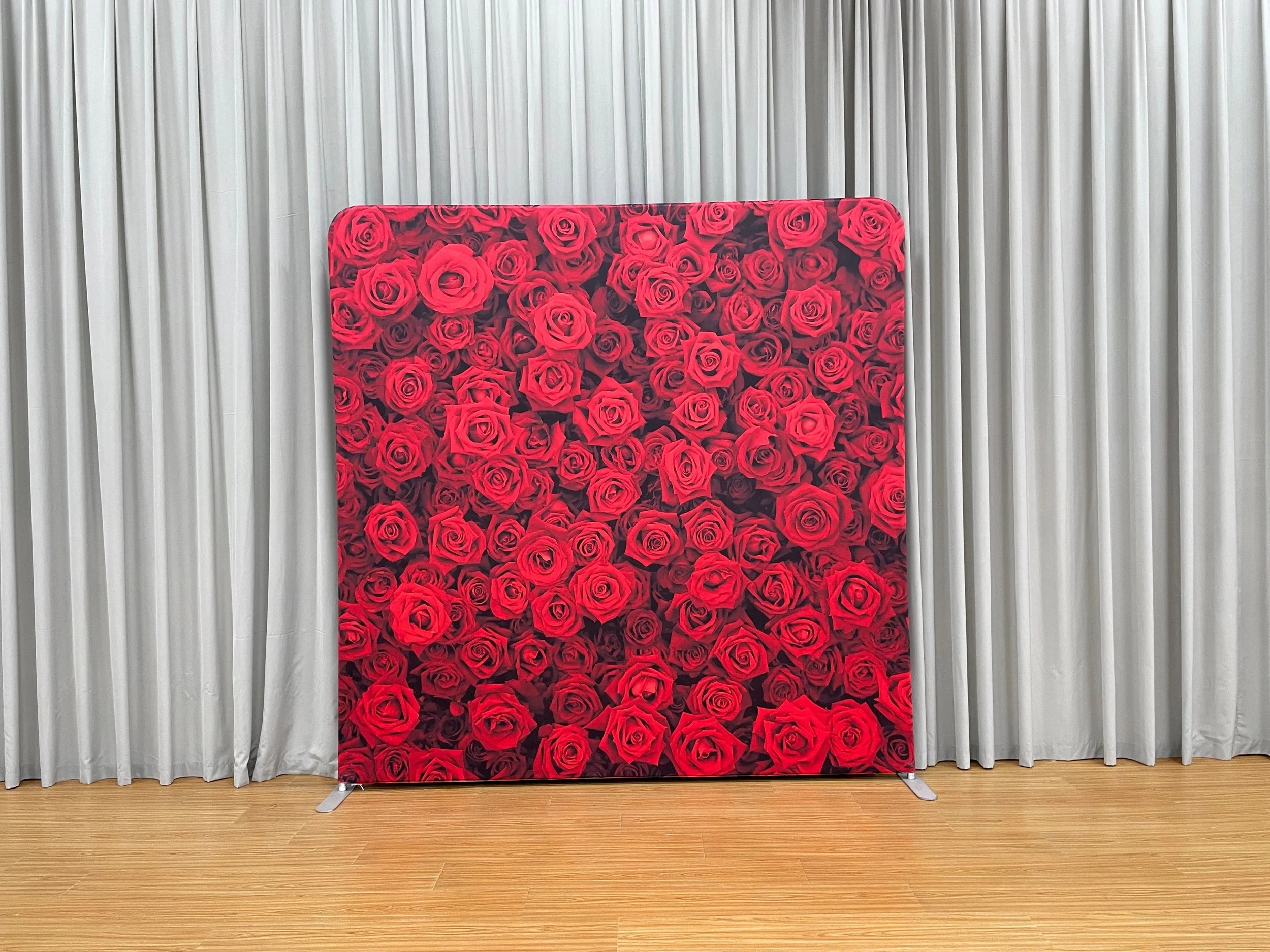 A decorative backdrop with a pattern of red roses stands in front of white curtains in a room with wooden flooring.