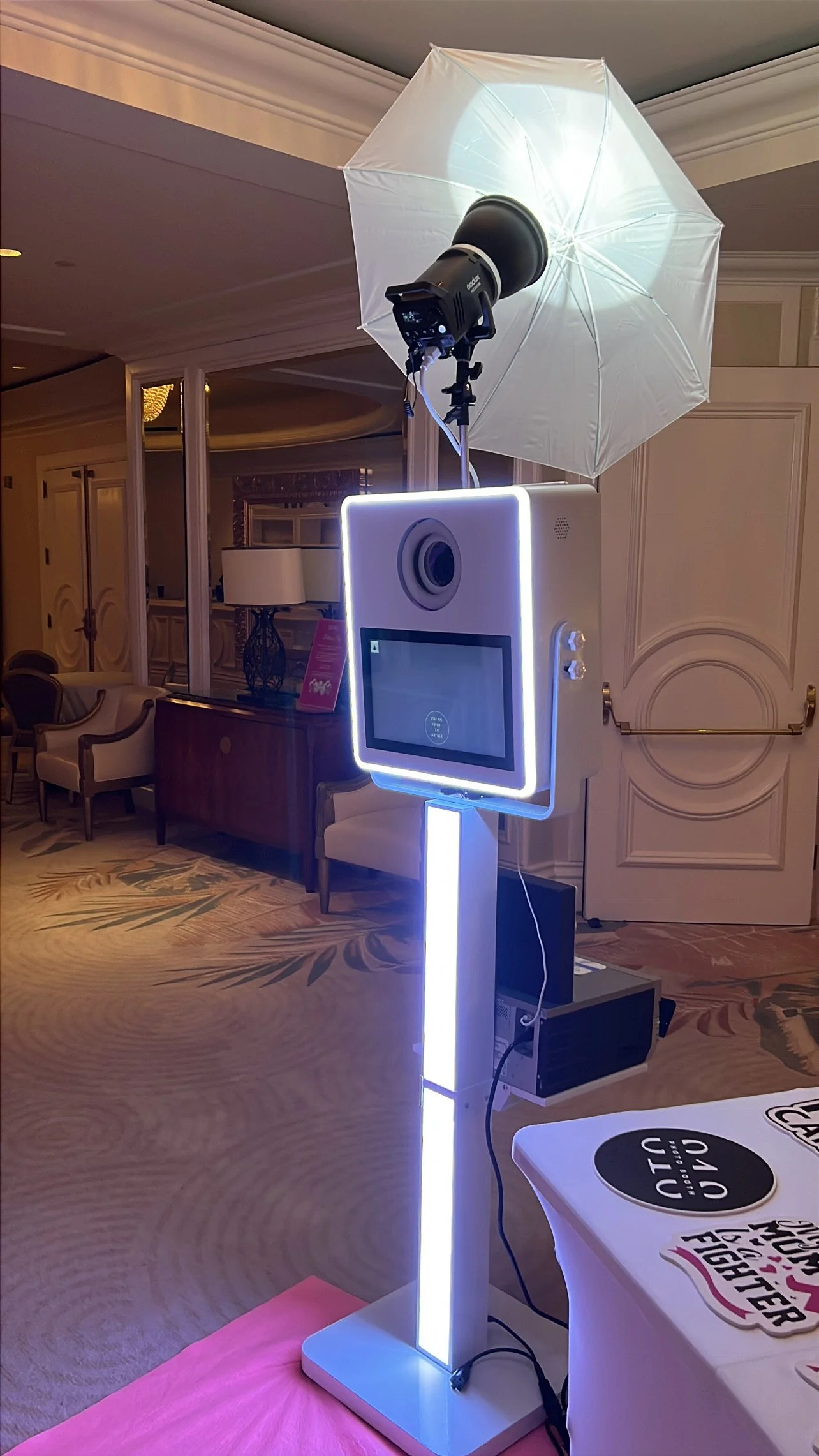 A photo booth with a large touchscreen, a camera, and a white umbrella light in an event space with elegant furniture.