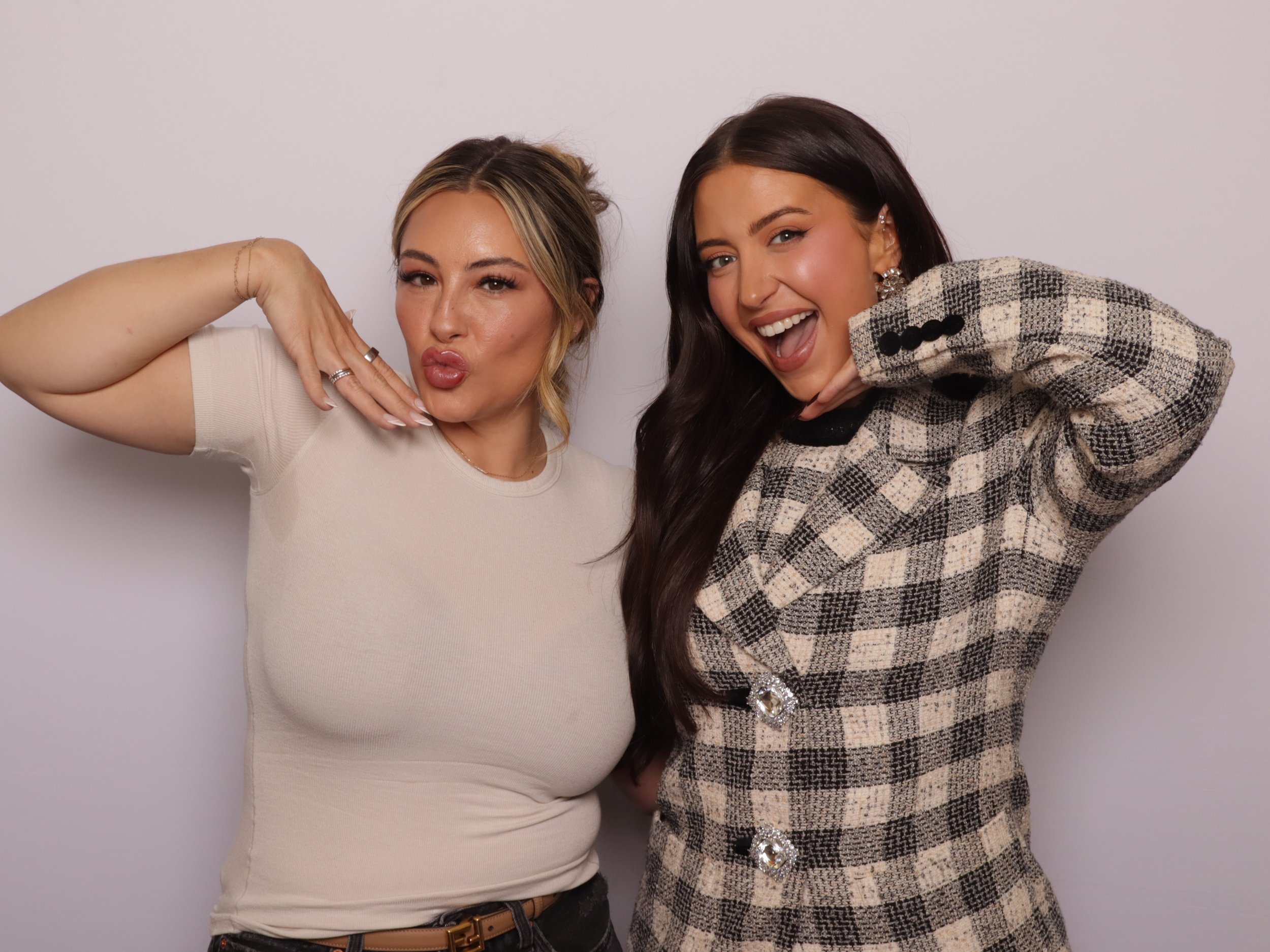 Two women posing playfully against a white background, one in a beige shirt making a kiss face and the other in a checkered blazer smiling with her hand on her chin.