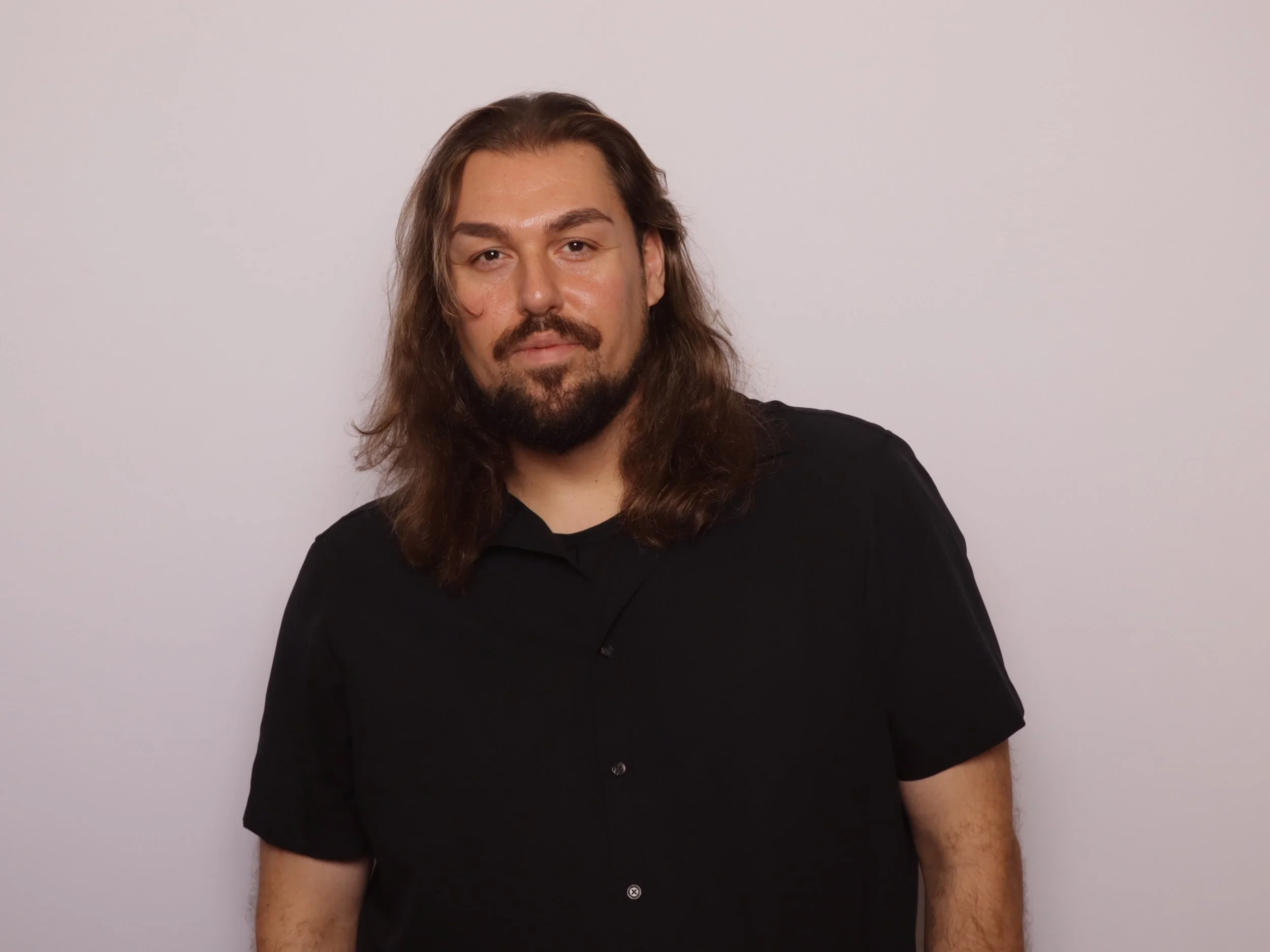 A man with long brown hair and a beard, wearing a black shirt, standing against a plain light-colored background.
