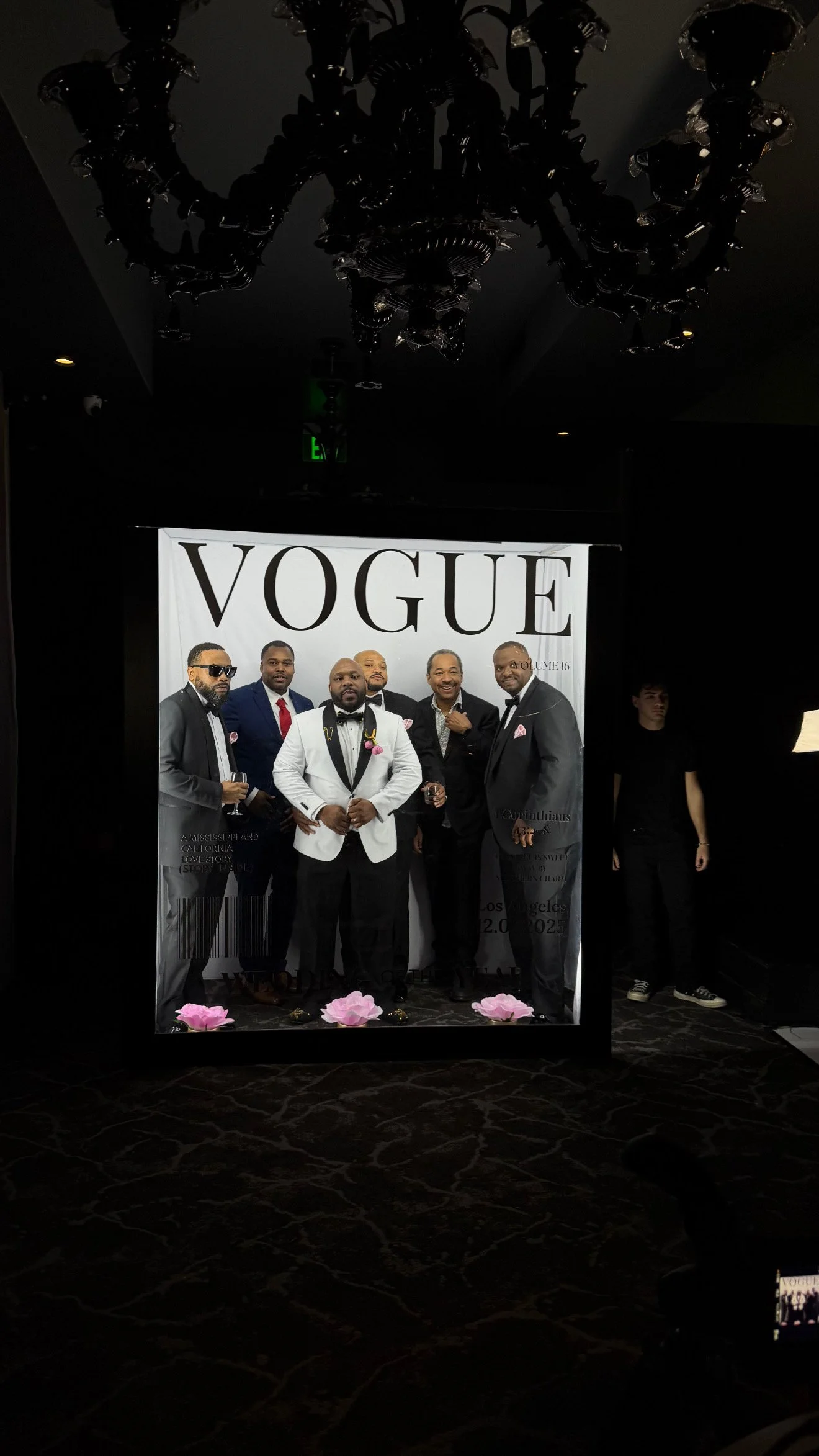Group of seven men in tuxedos posing in front of a large Vogue cover backdrop at an event, with two pink flowers on the floor in front of them.