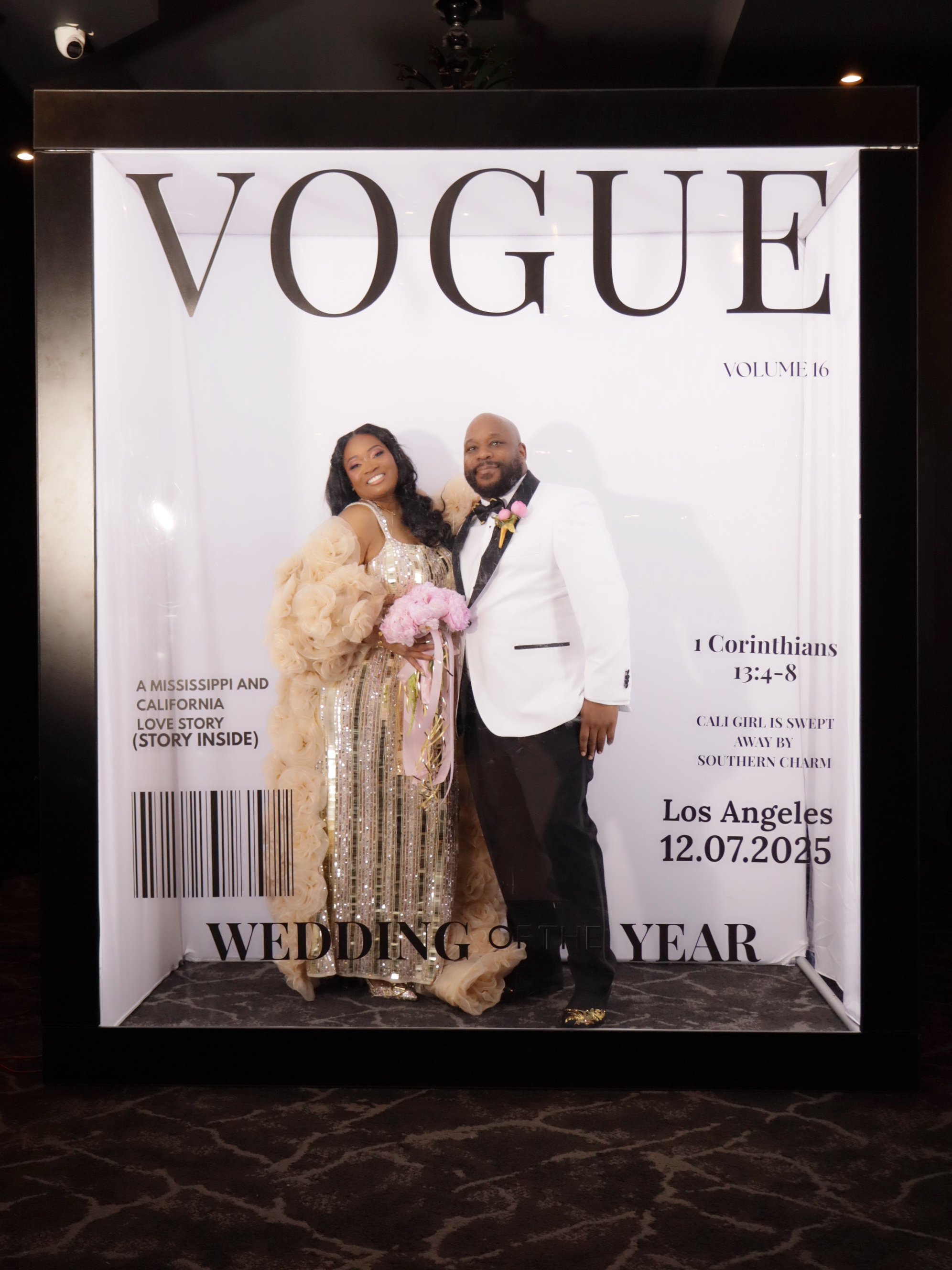 A couple dressed in formal attire standing in front of a large Vogue magazine cover backdrop at a wedding, with the bride holding a pink bouquet and wearing a gold sequined gown, and the groom in a white tuxedo jacket with a pink boutonniere.