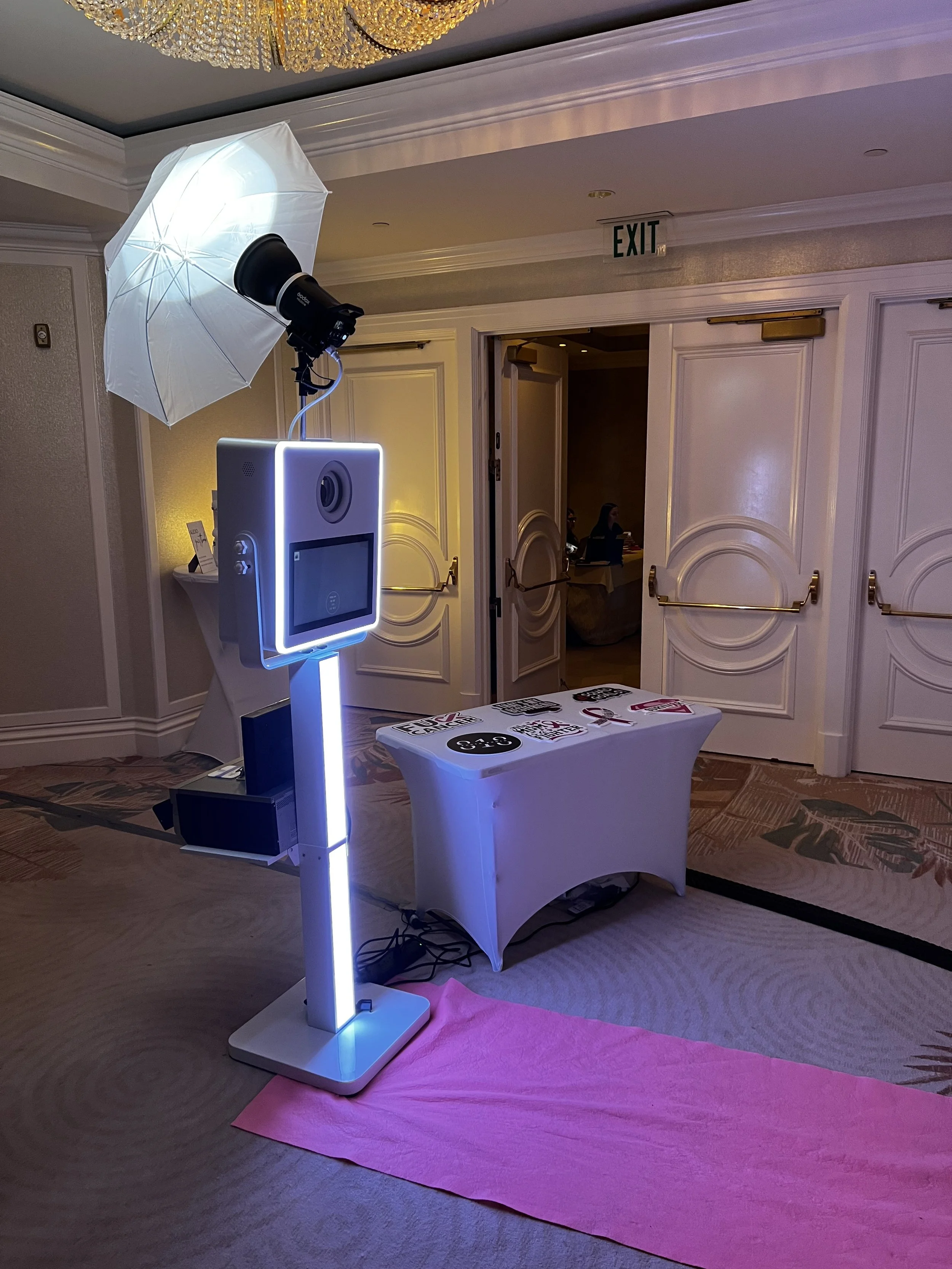 Photo booth setup with a camera, lighting umbrella, and a digital touchscreen, on a pink mat near a white table with photo props, in a hotel conference room.