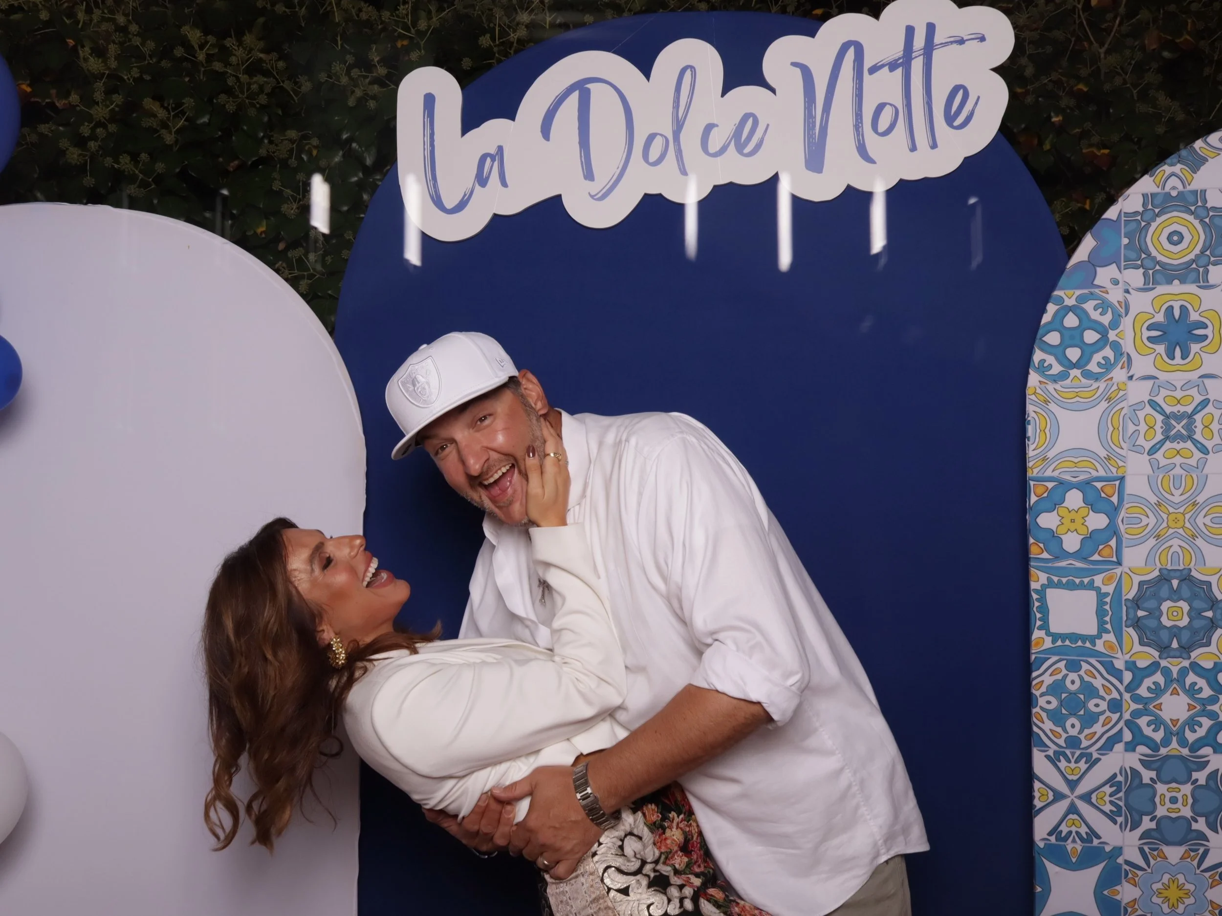 A smiling man and woman pose together in front of a colorful backdrop with the sign 'La Dolce Notte'. The man is wearing a white shirt and a white cap, while the woman is wearing a white top and floral skirt, and has long wavy hair.
