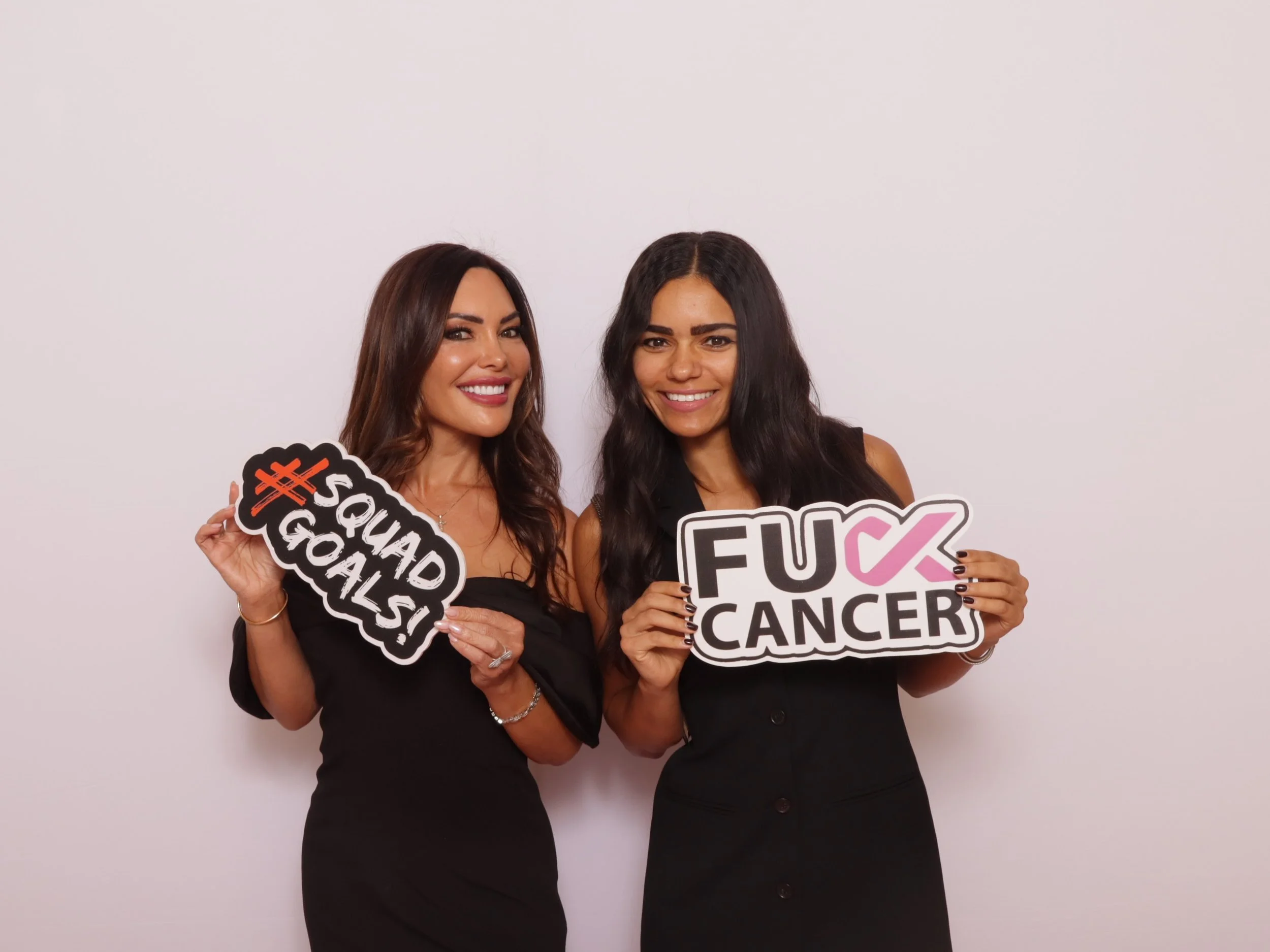 Two women smiling and holding signs supporting cancer awareness, one sign says '#SQUADGOALS' and the other says 'FUCK CANCER' with a pink ribbon.