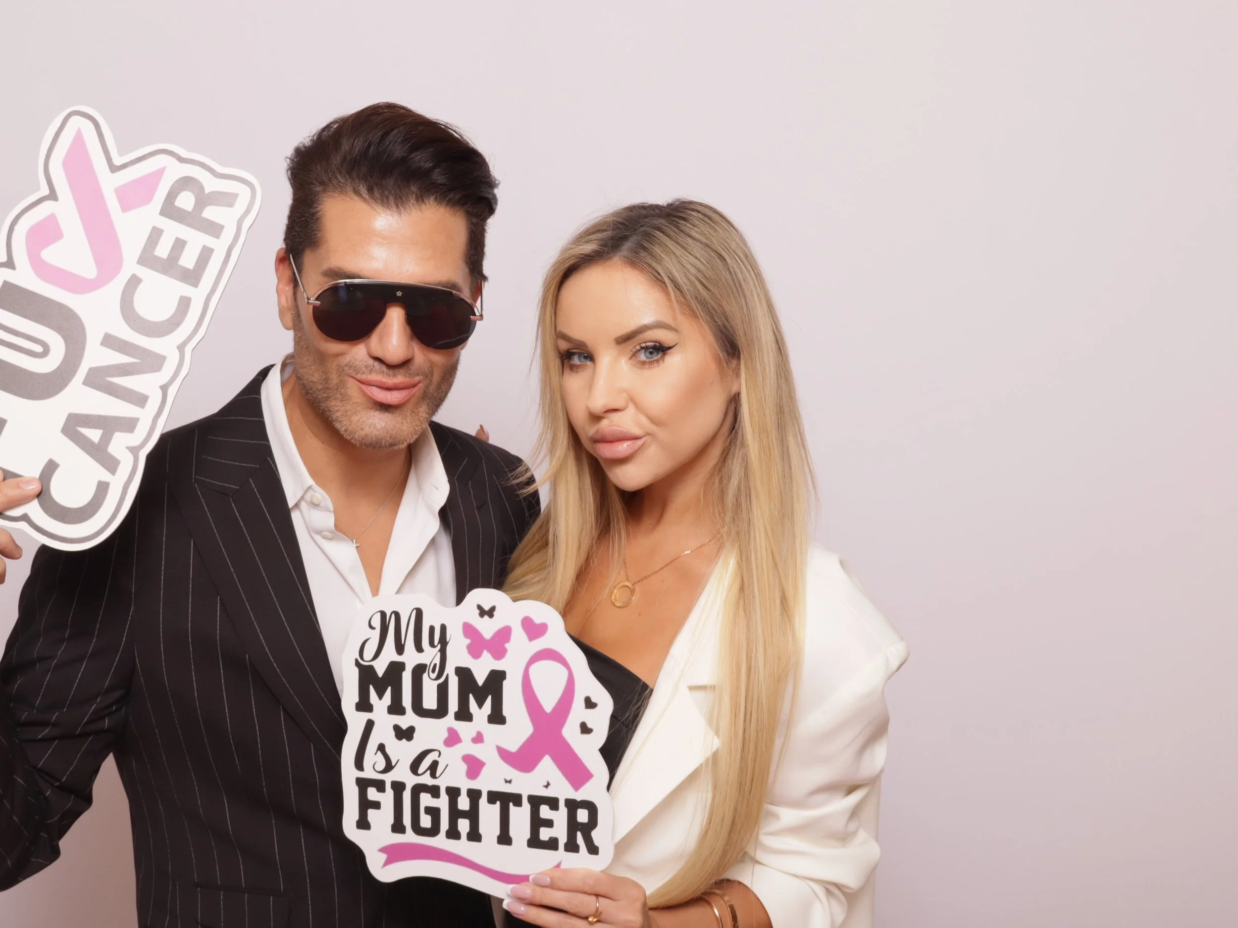 A man in a pinstripe suit with sunglasses and a woman in a white blazer holding signs for breast cancer awareness. The signs read "You Forever" and "My Mum is a Fighter" with pink ribbons and butterfly graphics.