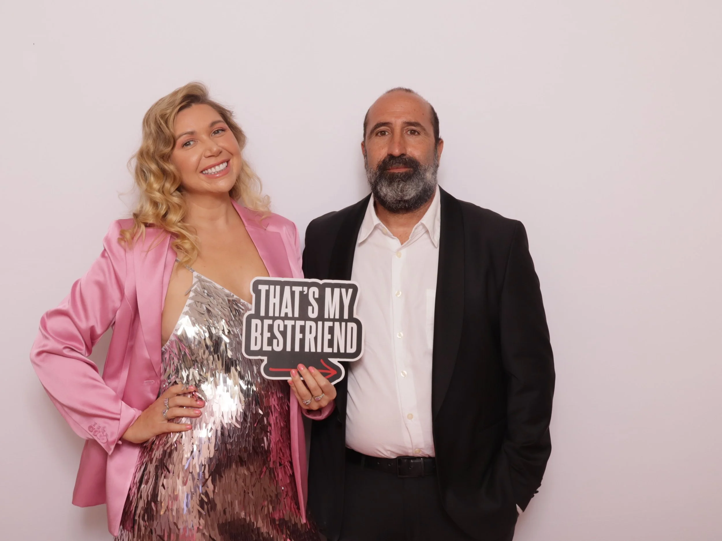 A woman and a man standing against a plain white background. The woman is holding a sign that says "That's my best friend." She is wearing a pink blazer and a shiny sequined dress, with blonde curly hair, smiling. The man has a beard, and is dressed 