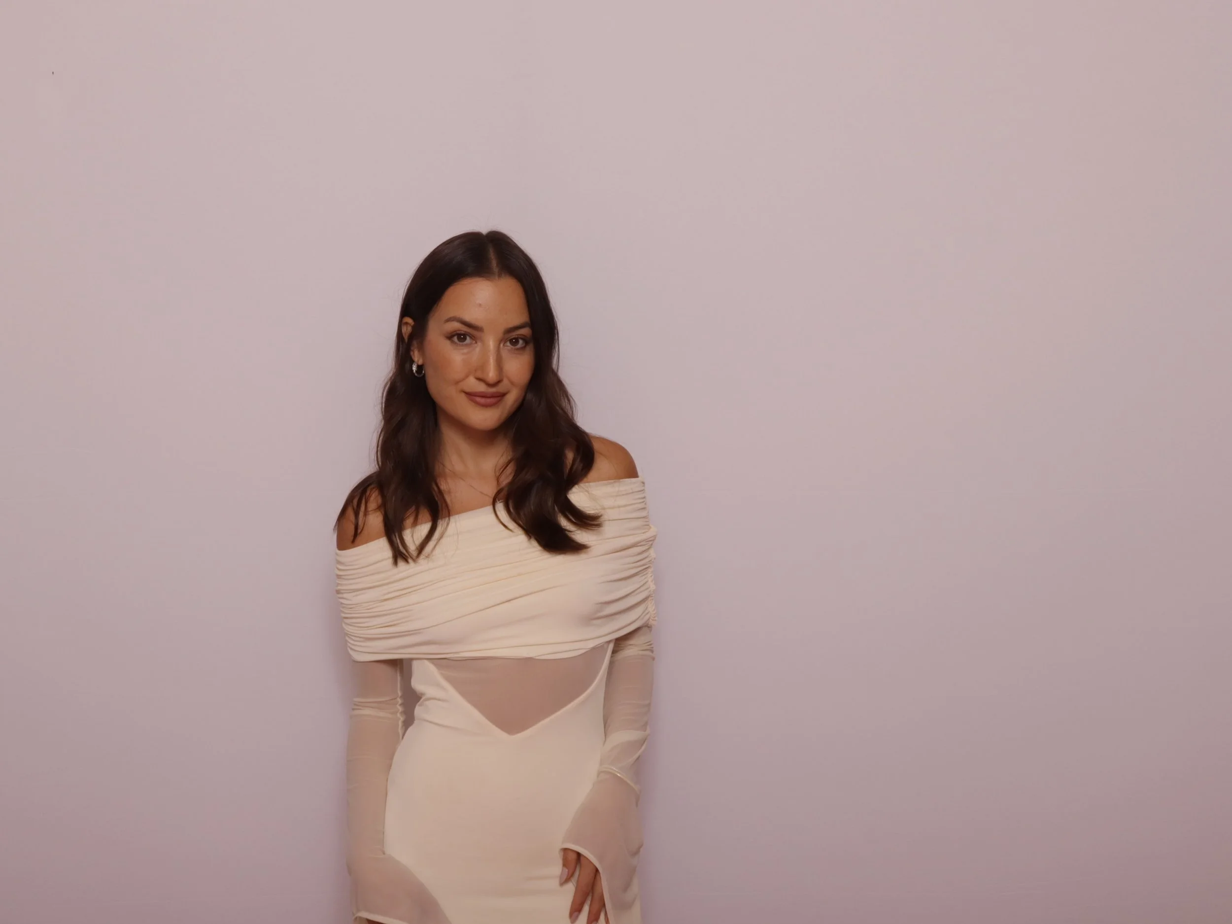 Woman with long dark hair in beige off-the-shoulder dress standing against plain white wall.