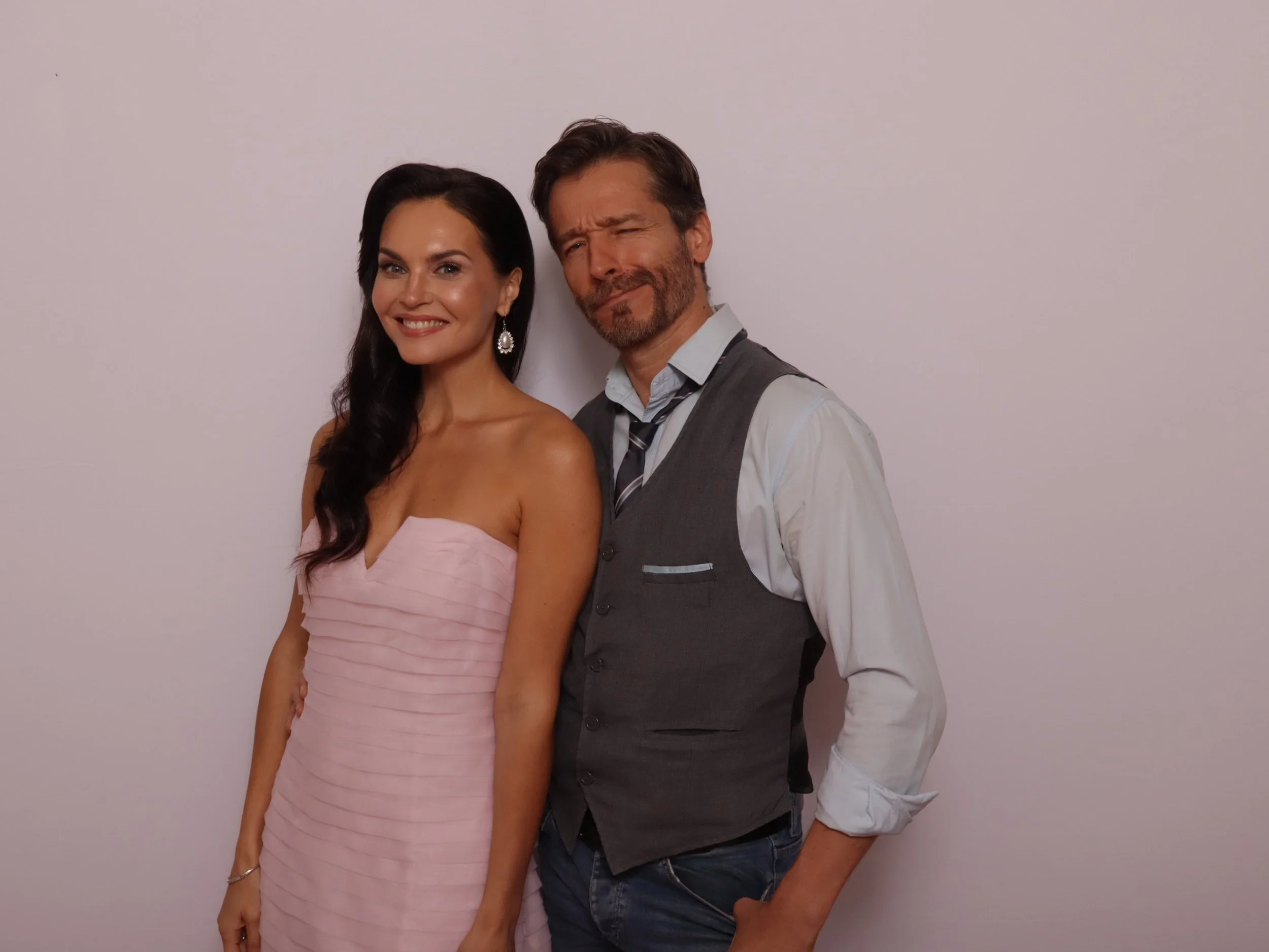 A woman in a pink strapless dress and a man in a gray vest and white shirt standing against a plain wall.