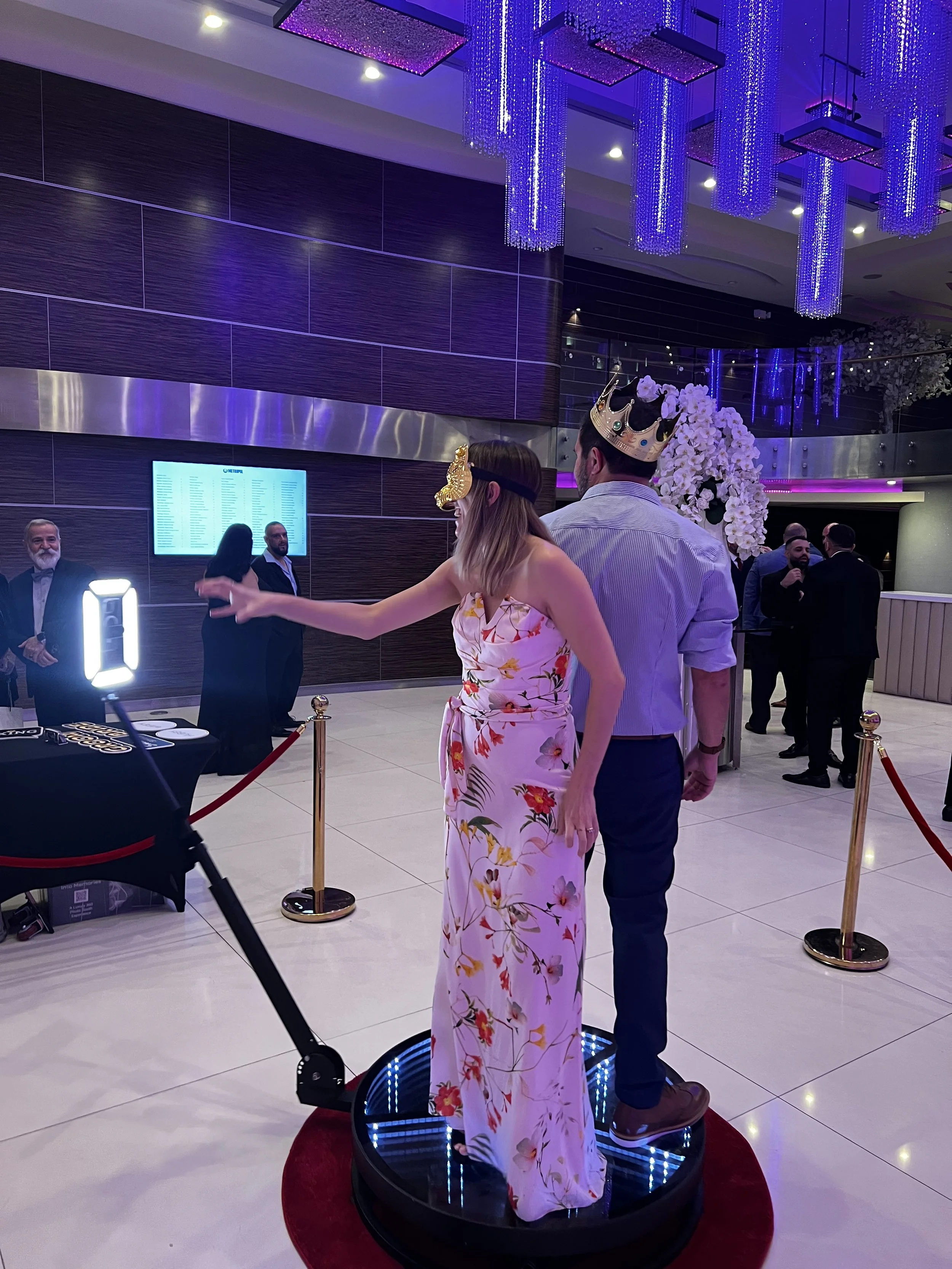 A woman and a man standing on a hoverboard, both wearing crowns, at a party or event with purple lighting, chandeliers, and floral decorations in the background.