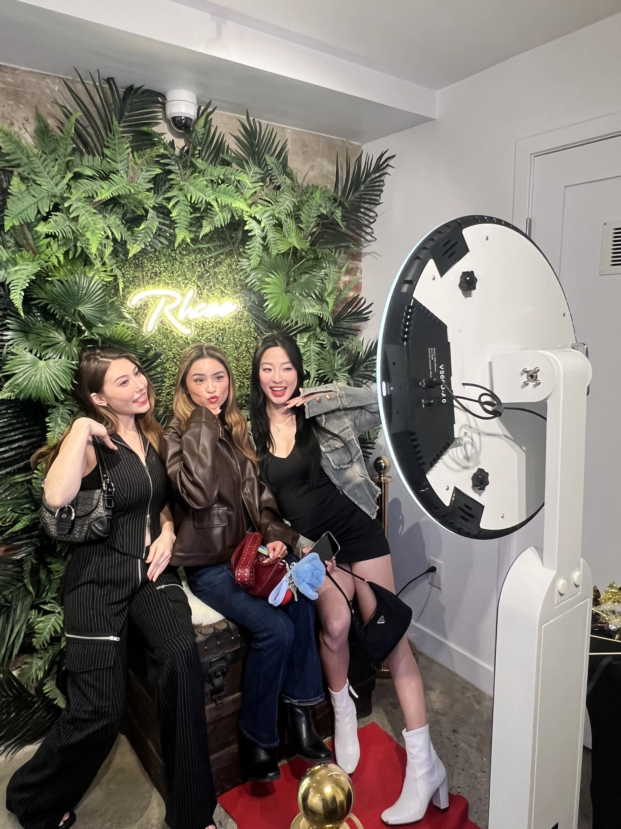 Three women posing for a photo in front of a green leafy wall with a neon sign that says 'Rhea'. A photo booth is set up nearby.