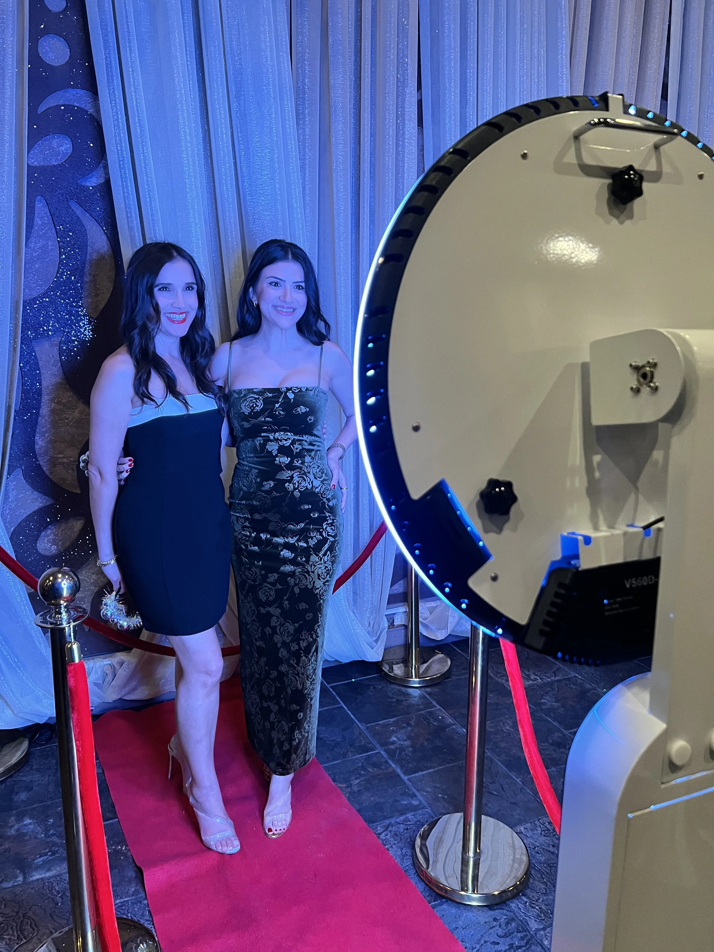 Two women in elegant dresses posing on a red carpet with a photo booth or photo station setup nearby.