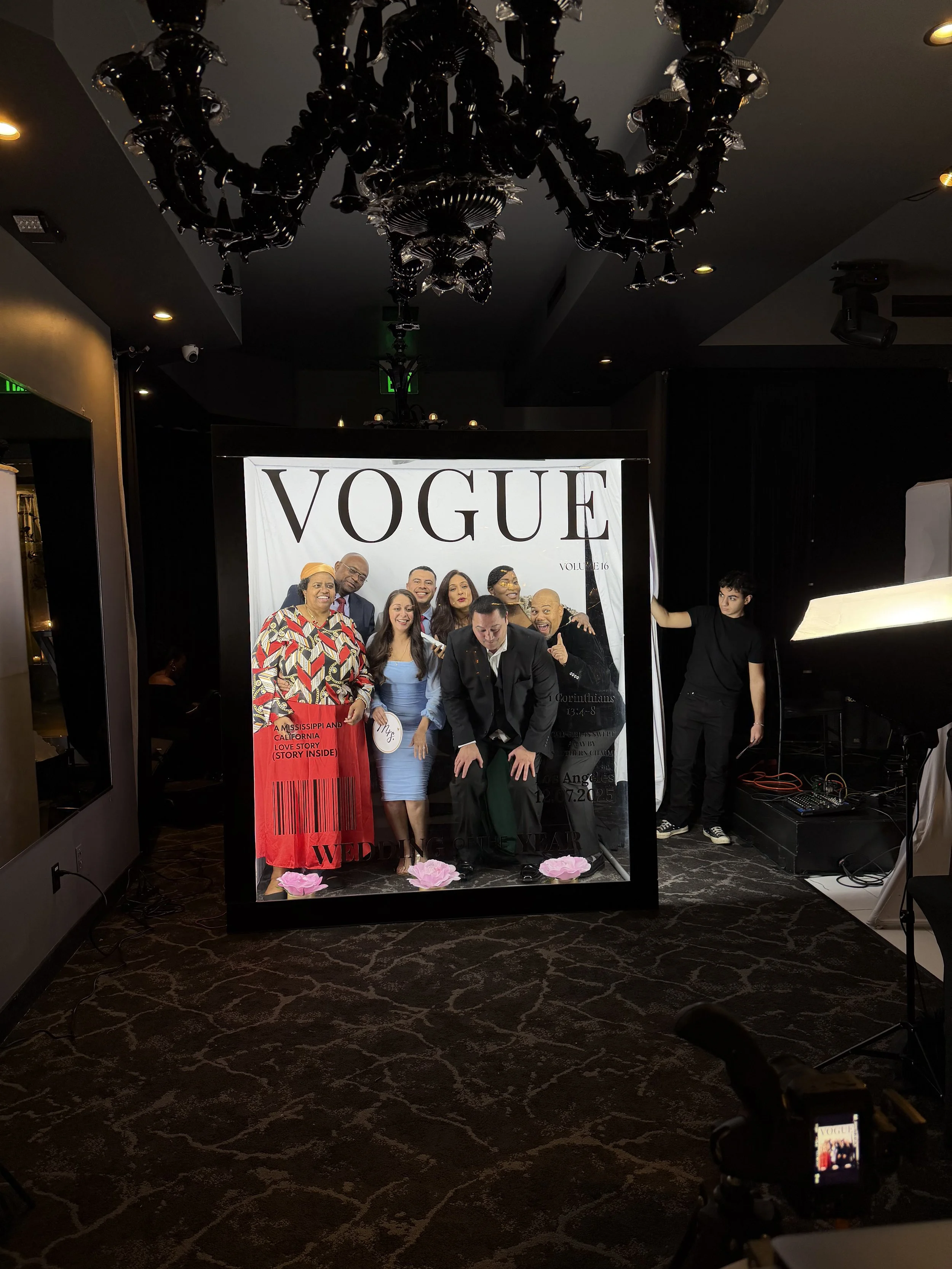 People posing for a photo in a photo booth with a Vogue magazine cover style backdrop. The group includes men and women dressed in formal attire, and a young man to the side adjusting equipment in a dimly lit room with a chandelier overhead.