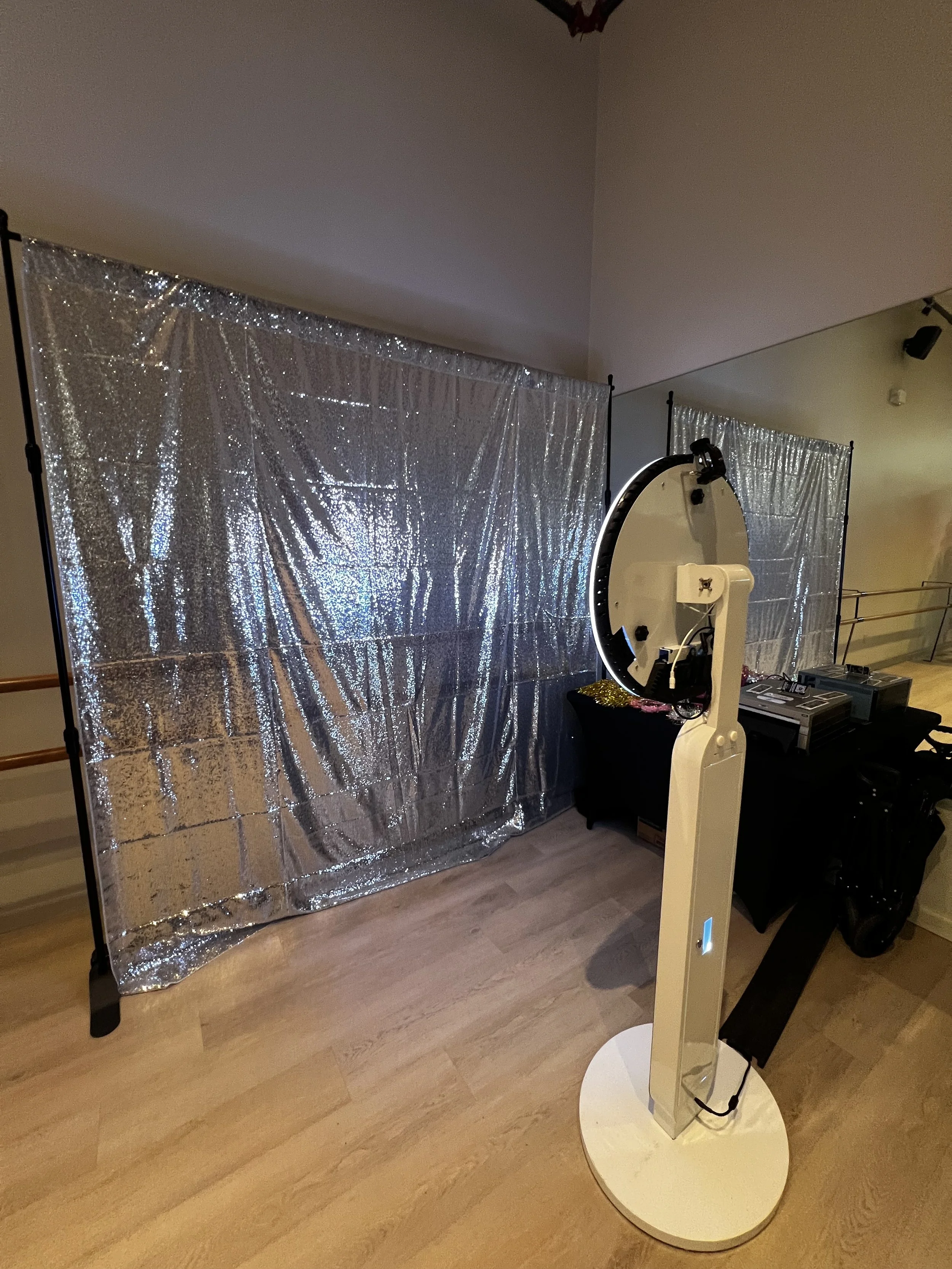 A photo booth setup with a shiny silver backdrop, a standing mirror photo booth, and some equipment on a table.