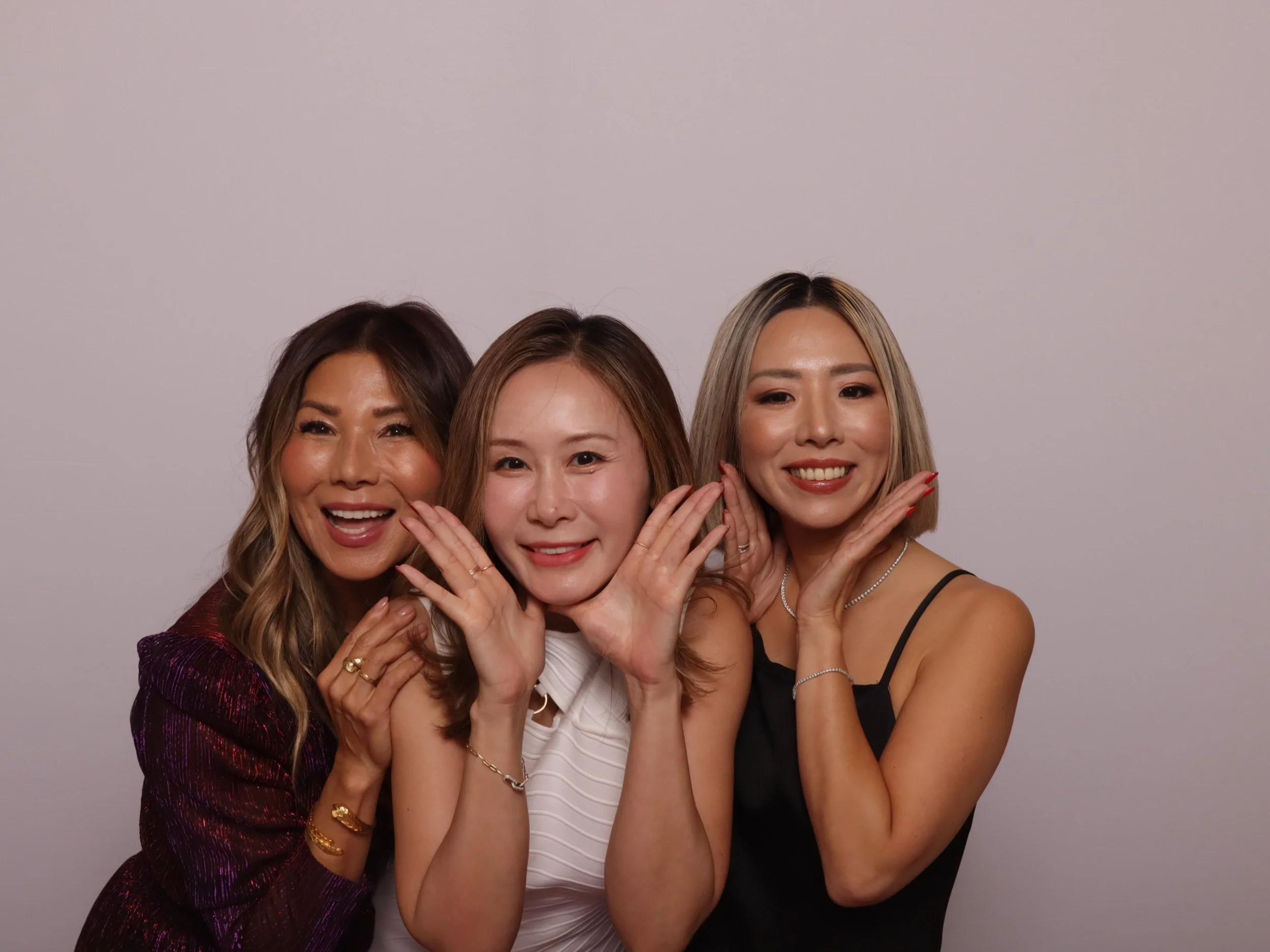 Three women smiling with hands framing their faces against a plain background.