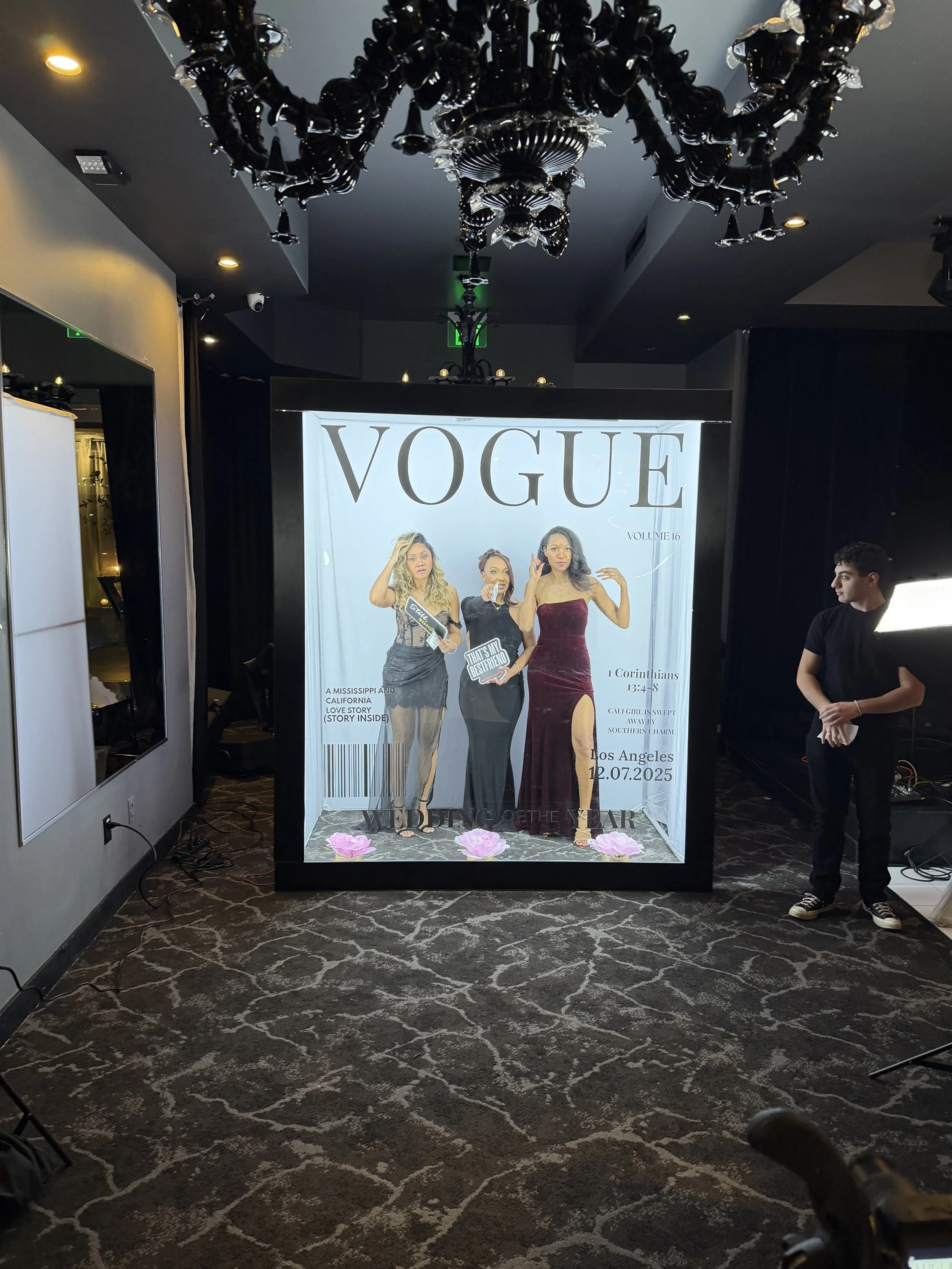 Life-sized mock magazine cover resembling Vogue featuring three women in glamorous dresses, with text and decorative elements, set inside an event space or hotel suite.