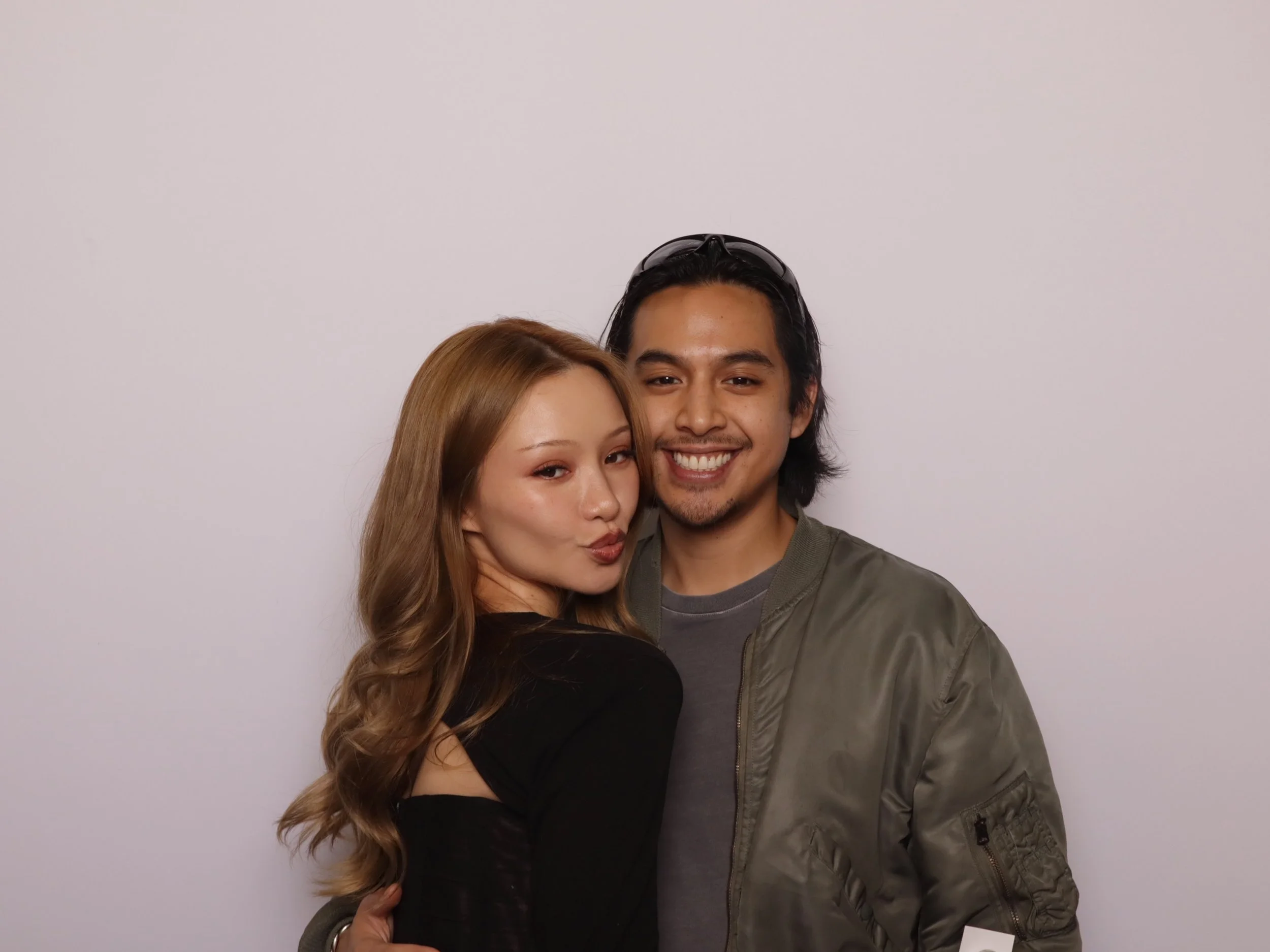 A young woman and man posing together, smiling at the camera against a plain light-colored background.