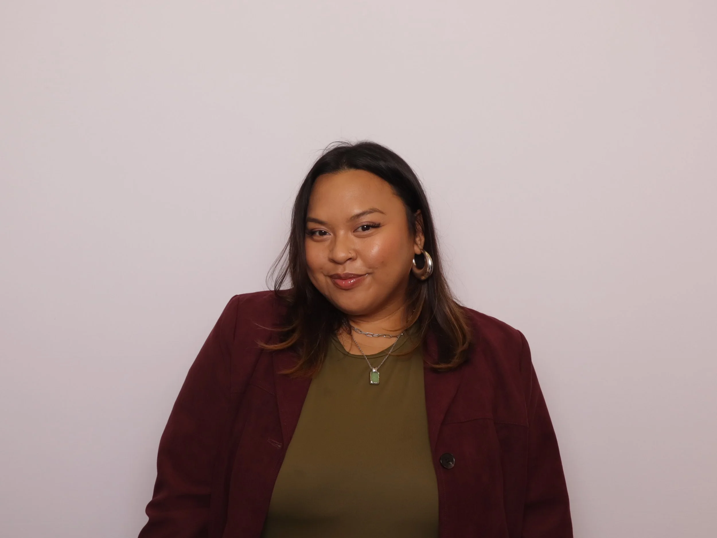 A woman with shoulder-length dark hair wearing a green top and a maroon jacket, accessorized with hoop earrings and layered necklaces, smiling softly against a plain white background.