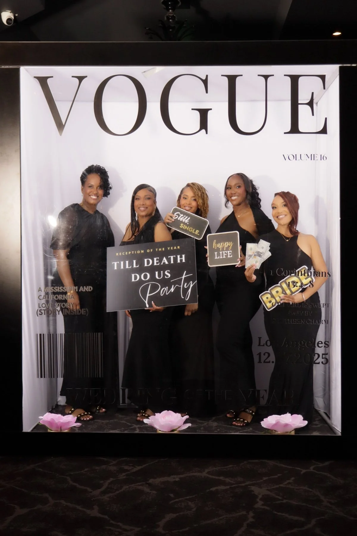 Group of five women posing in front of a Vogue magazine-themed photo backdrop, holding signs and fake money, dressed in black with pink flower decorations at their feet.