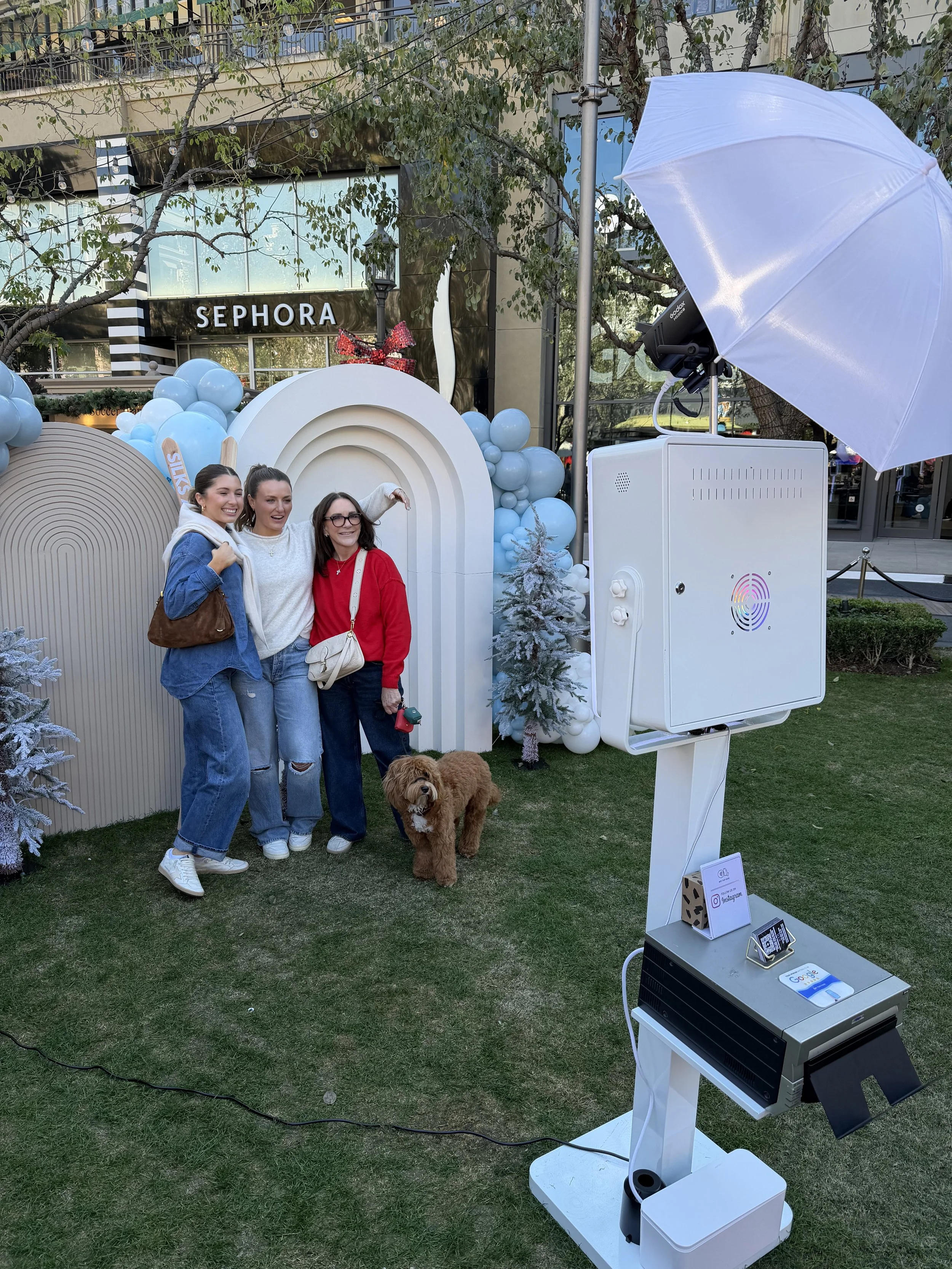Three women and a dog posing for a photo outdoors with an interview setup, including a white umbrella, sound equipment, and decorative balloons and trees in the background.