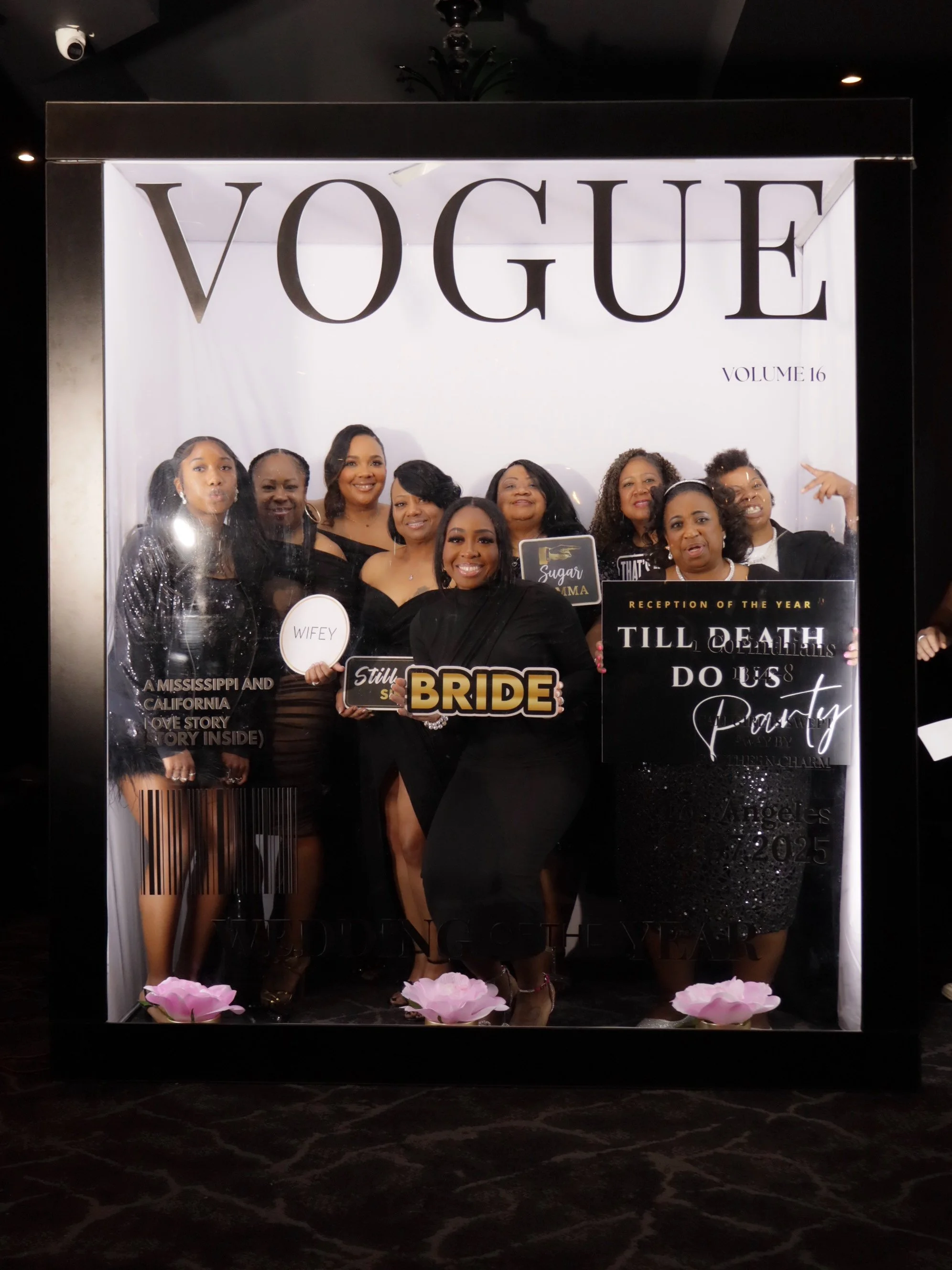 A group of women posing together at a formal event, holding signs including 'BRIDE' and 'WIFEY', with a backdrop displaying 'VOGUE' and various text elements.