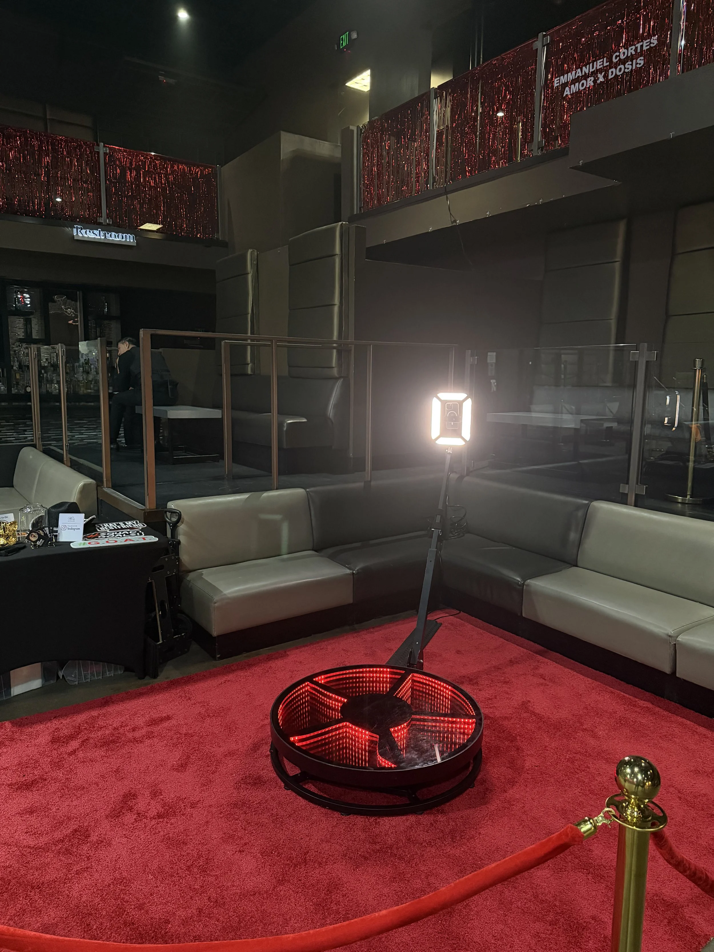 A lit pool table in front of a beige sofa and a gold stanchion with red velvet rope in a dimly lit entertainment or lounge area.