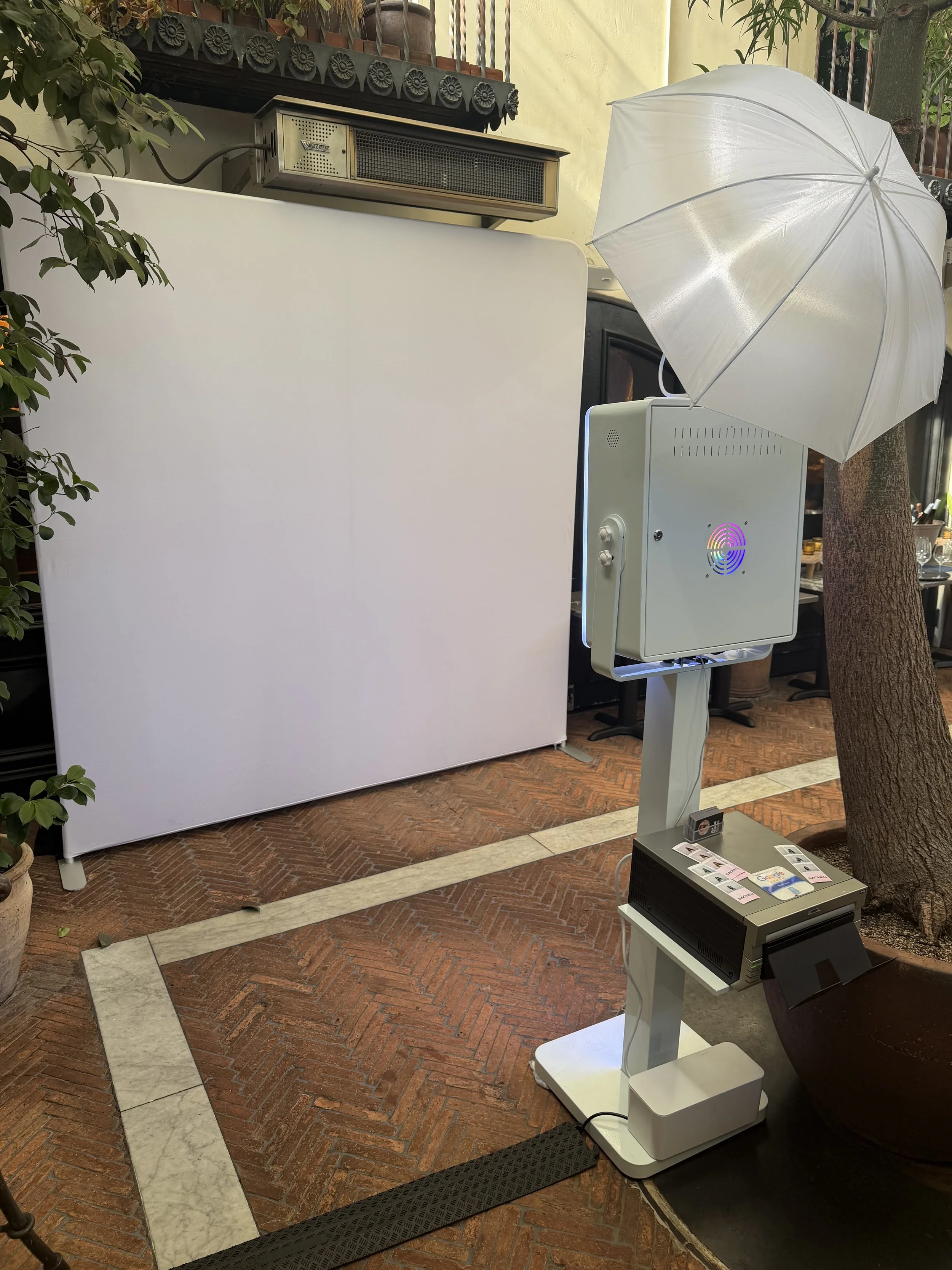 Photo of a photo booth setup with a large white backdrop, an umbrella for lighting, and a touchscreen kiosk, situated outdoors on a brick-paved area near a tree and potted plants.