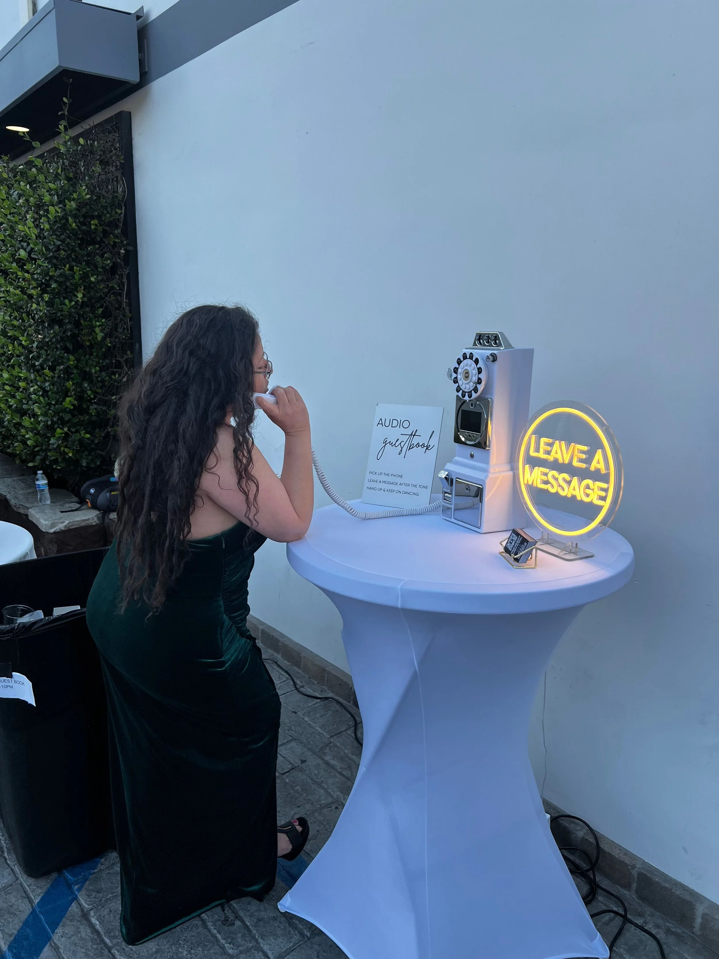 A young woman with long curly hair in a black dress uses a payphone at a wedding guestbook station. The station includes a vintage phone booth with a rotary dial, a neon sign that reads 'Leave a Message,' and a sign instructing to pick up the phone a
