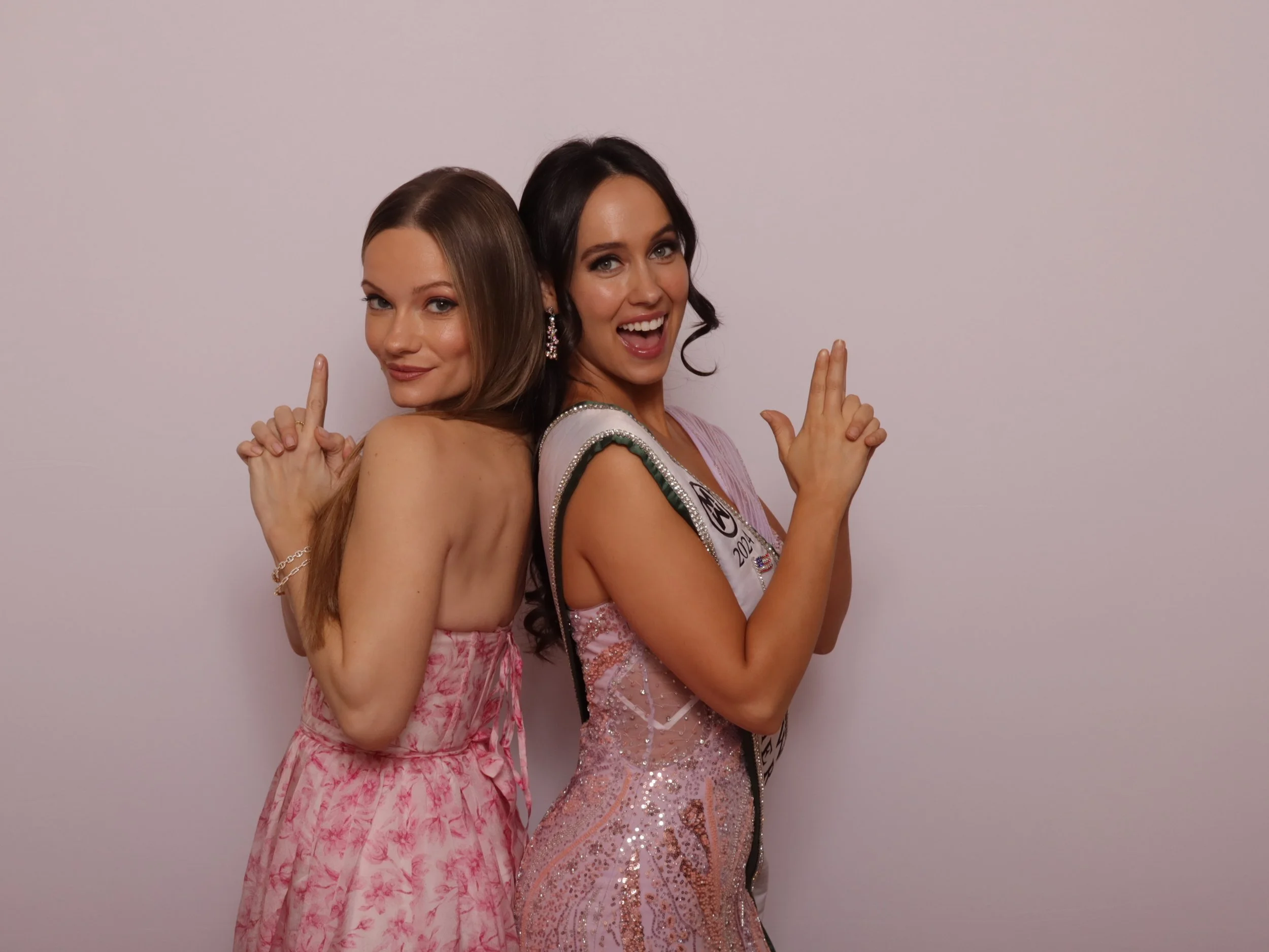 Two women in elegant dresses, back to back, smiling and showing gesture with their hands against a plain background.